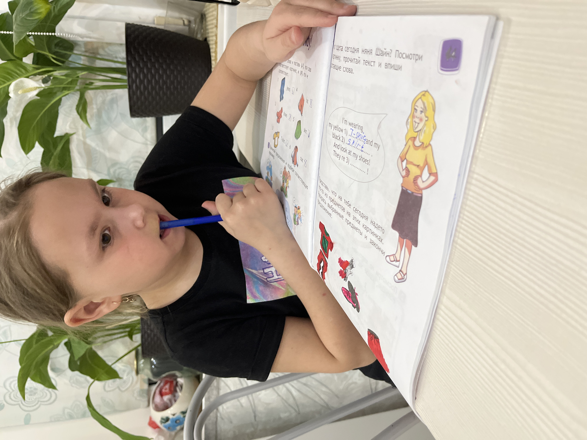 A young girl is pictured indoors, likely at a home desk or table in Chaikovsky, Russia, engrossed in a textbook or workbook. She is seen from a slightly elevated angle, with a blue pen held to her mouth, indicating concentration. Her right hand rests on the open book, which lies on a light-colored, ridged surface.
The workbook displays educational content, primarily an English language exercise. Visible English text includes "I'm wearing my yellow 1)...Shand my black 2) S_k_ir_t..... And look at my shoes! They're 3)............!", accompanied by a cartoon drawing of a smiling woman. There are also images of clothing items like "jeans," "shoes," "T-shirt," "hat," "skirt," and "coat," with checkboxes. Russian instructions on the page translate to: "What is Nanny Shine wearing today? Look at the picture, read the text and fill in the missing words," and an activity to "Imagine what you are wearing today from these items in these pictures. Describe the selected items and finish the sentence."
In the background, a lush green peace lily plant in a black woven pot is visible. The wall behind it features patterned wallpaper. A decorative skull-shaped item with blue floral details is partially seen on the table. The indoor lighting suggests the activity could be taking place during the day or evening. The girl's focused demeanor and the academic materials indicate she is engaged in learning or homework.