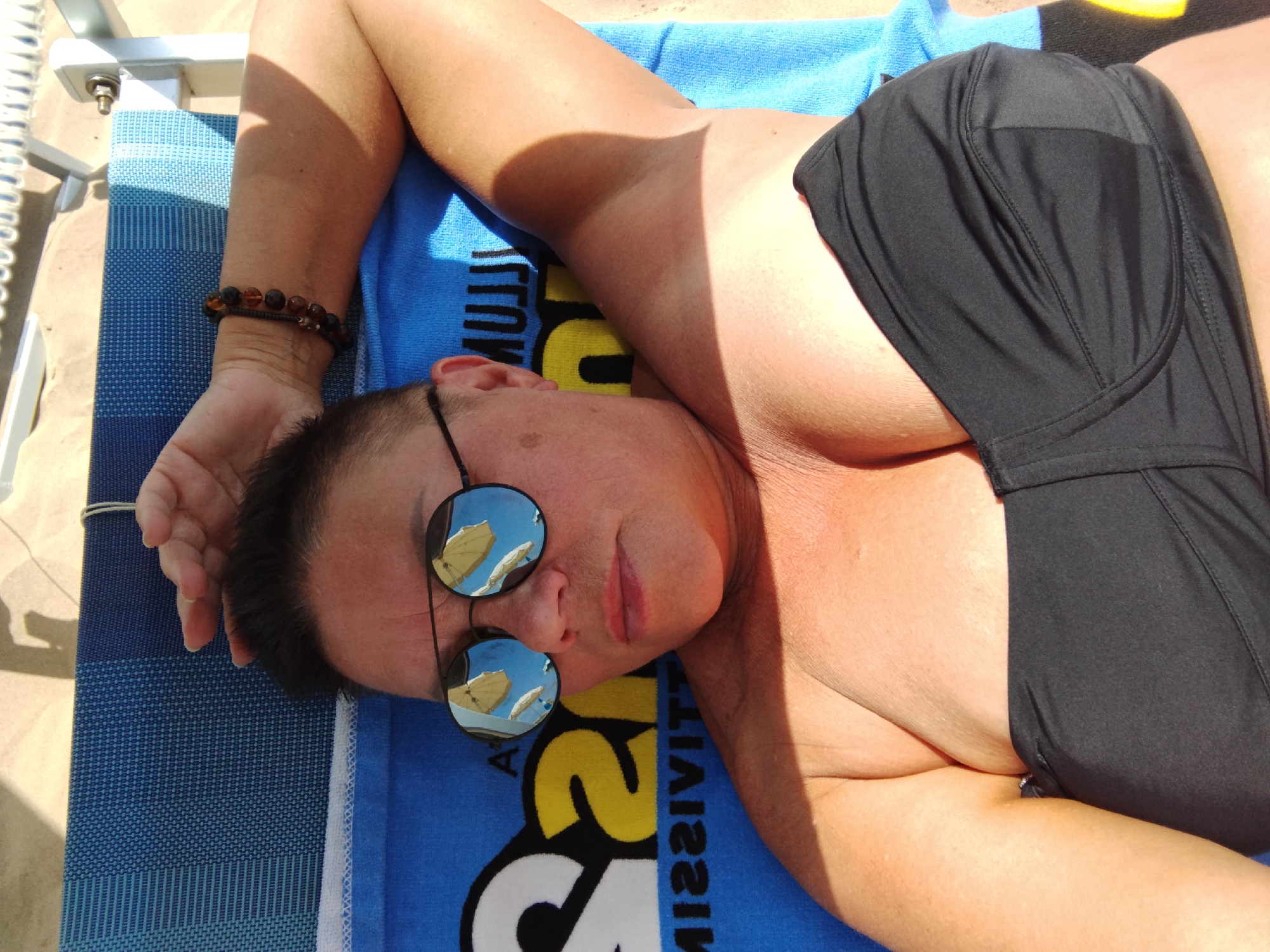 An overhead shot shows a person lying on a blue and white striped beach chair on a sandy beach. The person is wearing a black bikini top and sunglasses. The sunglasses have round lenses that reflect the beach scene, showing beach umbrellas. The person's arm is bent and resting on their head, and their other arm is resting by their side. The background is blurred, but appears to be a beach with people and umbrellas. The lighting suggests it is a sunny day. The image was taken in Peschici, Italy.