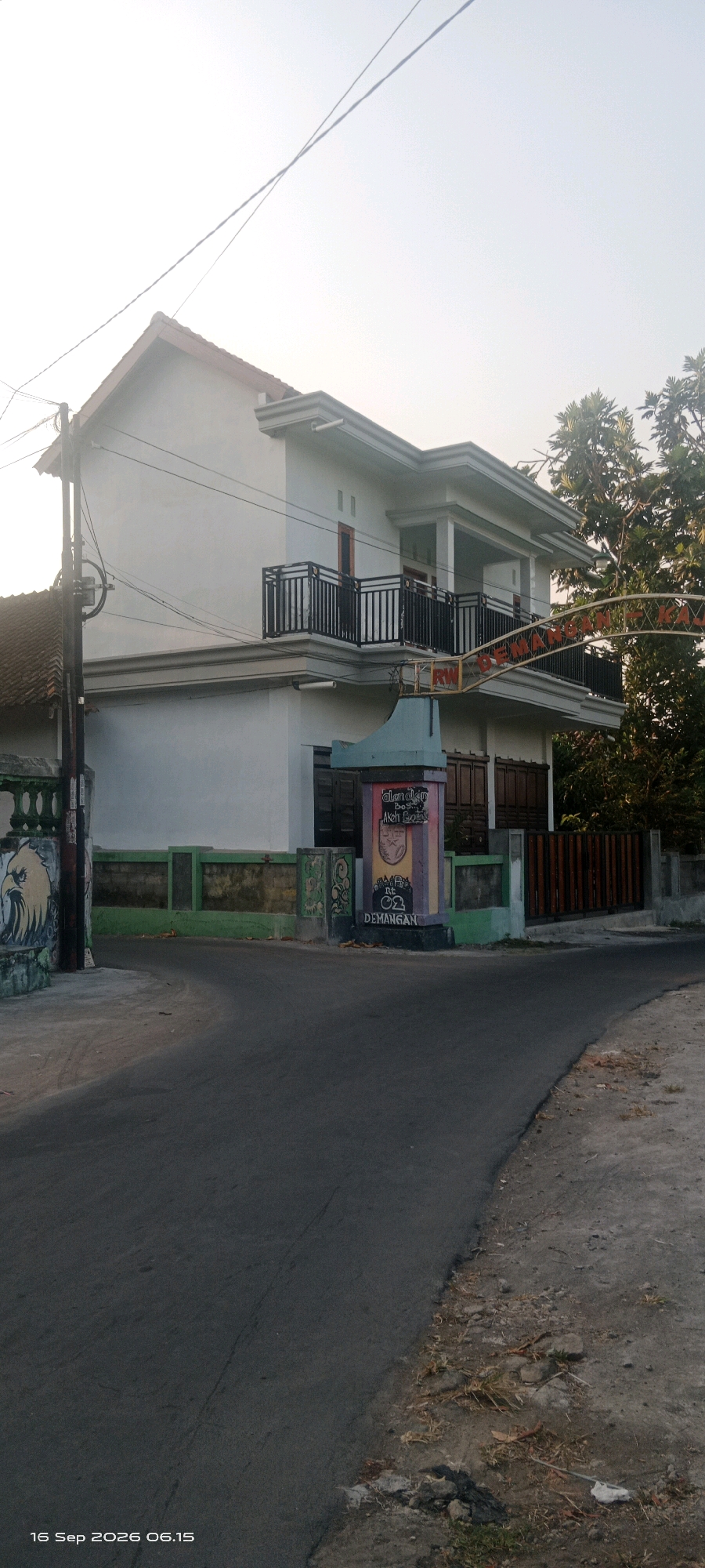 In Glodogan, Indonesia, this image captures a two-story house on a corner lot beside a paved road. The house, painted white, has a red-tiled roof and black metal railings on its balconies. A prominent decorative pillar stands on the right side of the road, featuring colorful graffiti and the text "DEMANGAN RW 02". To the left of the pillar, a green fence with decorative elements runs along the property line. Further to the left, a mural of an eagle's head is visible on a wall. Trees line the right side of the road behind the house. The scene is illuminated by soft sunlight, suggesting it was taken in the morning or late afternoon. The timestamp in the bottom left corner indicates the image was captured on September 16, 2026, at 06:15.