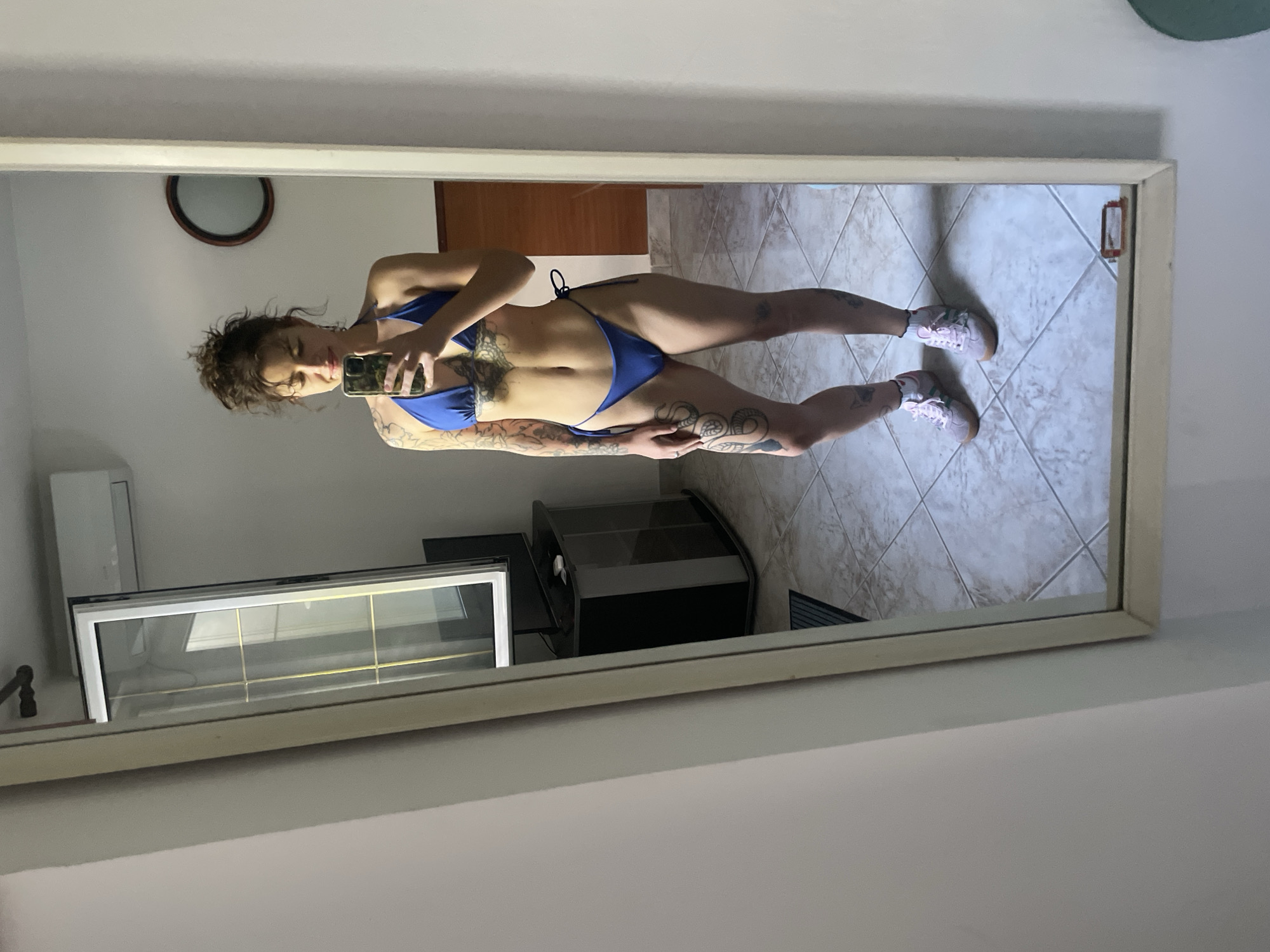 A young woman with tattoos is taking a mirror selfie in a room. She is wearing a blue bikini and pink sneakers. The room appears to be a bedroom or living area, with tiled floors, a mirror, a television stand, and a window. The window offers a view of an outdoor area, possibly a balcony or street. The overall lighting suggests it is daytime.