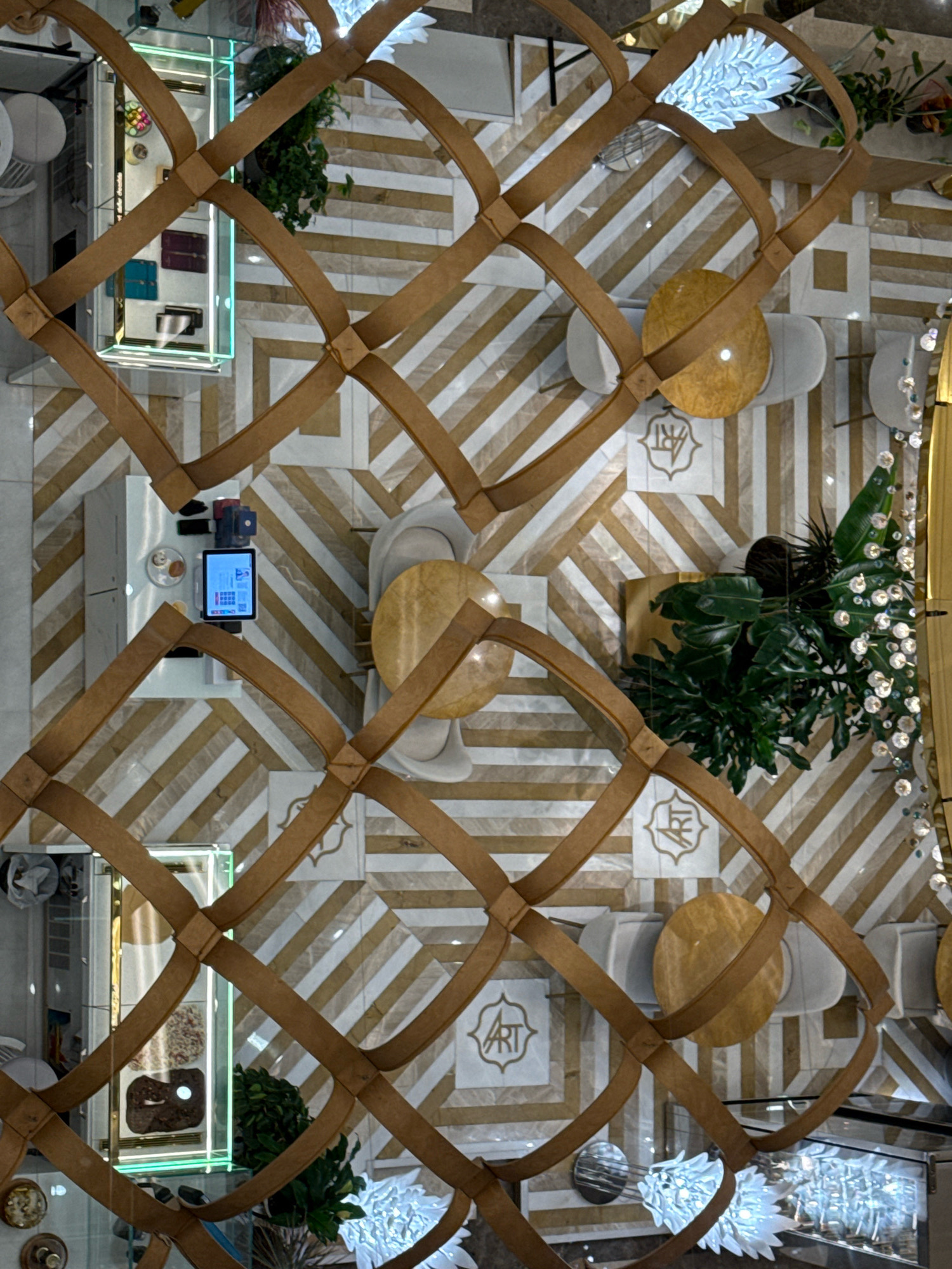 The image provides a high-angle, overhead view of an indoor space, possibly a cafe or retail establishment, featuring a decorative brown lattice structure in the foreground that partially obscures the scene below. The floor is covered with a geometric pattern of alternating white and gold diagonal stripes, creating a visually dynamic surface. Scattered throughout the space are several round, golden-colored tables, some accompanied by white seating. Decorative elements include lush green plants in pots, and abstract white light fixtures that resemble floral arrangements. A few display cases with products, a tablet device, and what appear to be food items are also visible. The overall aesthetic suggests a modern and stylish interior. The location is specified as Tashkent, Uzbekistan, though no specific landmarks or identifying features are explicitly visible in the image to confirm this. There are no people present in the visible area of the image.