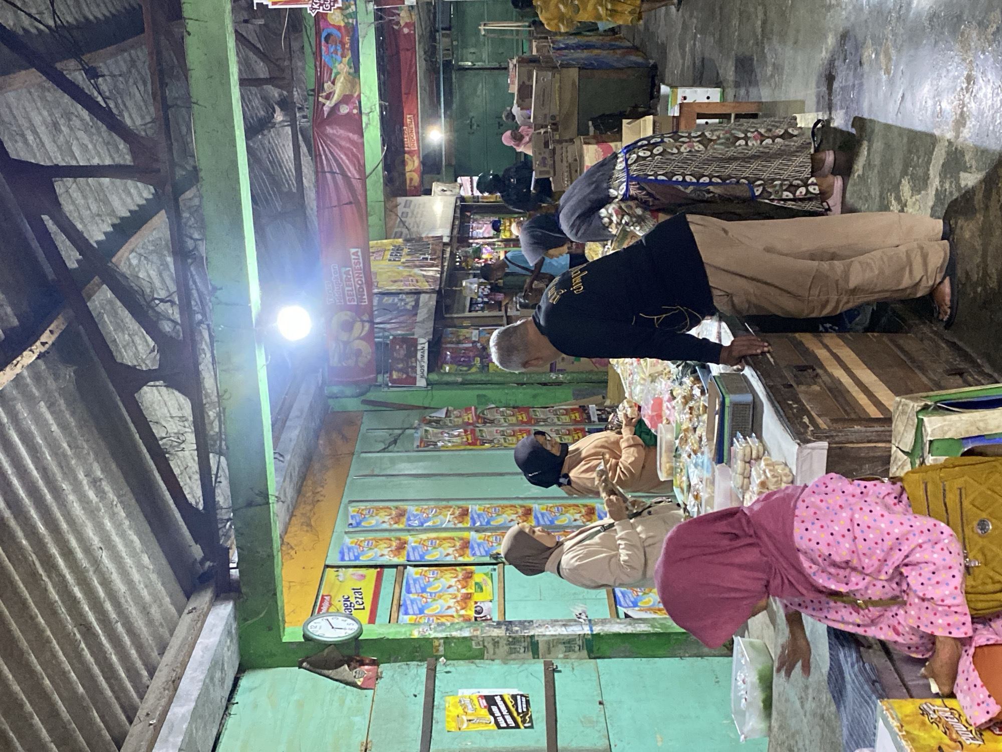In Ambarketawang, Indonesia, a dimly lit market stall displays goods under a corrugated metal roof supported by wooden beams. The stall is painted a vibrant green, with yellow accents along the ceiling. Various products are visible, including packaged drinks and snacks. Several individuals are present: a woman in a pink headscarf and polka-dot dress sits at the front, facing away from the camera, with a yellow bag beside her. Two women stand at the counter, one in a light brown hijab and the other in a dark hijab, both appearing to be customers. A man in a black t-shirt and tan pants is visible from the back, leaning on the counter. Further back, other individuals are browsing or working. The ground is concrete and appears wet in places, reflecting the light from overhead bulbs. Banners advertising products are hung above the stalls.