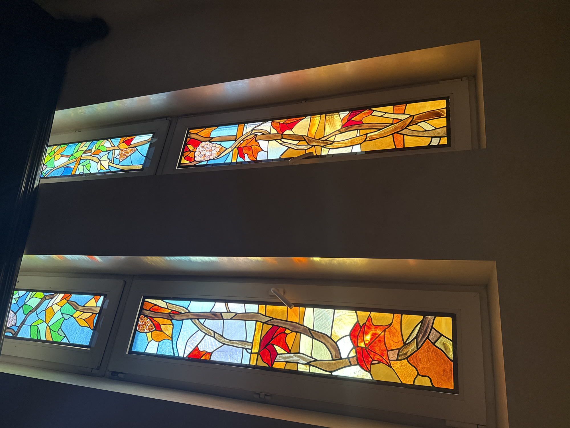 The image shows two tall, narrow stained-glass windows in what appears to be a building. Each window is divided into two vertical sections. The stained glass depicts abstract patterns with organic shapes, predominantly in shades of blue, green, yellow, orange, and red, resembling leaves and branches. Sunlight streams through the windows, casting colorful patterns of light on the adjacent wall. The left window has a white frame and a visible handle on its lower section. The right window also has a white frame and is positioned slightly further away. The walls appear to be painted a neutral, muted color. The overall impression is one of warm, natural light filtering through decorative glass. There is no specific information available to identify this location within the city of Yukki, Russia.