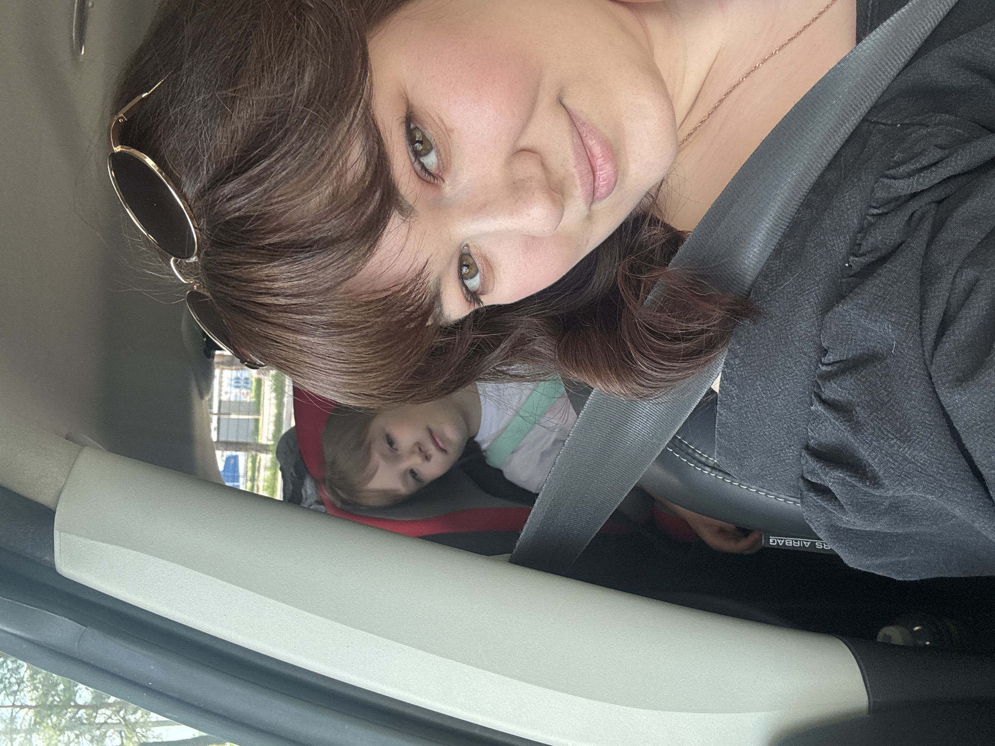 A woman is seated in a car wearing a seatbelt and sunglasses propped on her head. A child in a car seat is visible behind her. The location is not identifiable as Chaikovsky, Russia based on the image.
