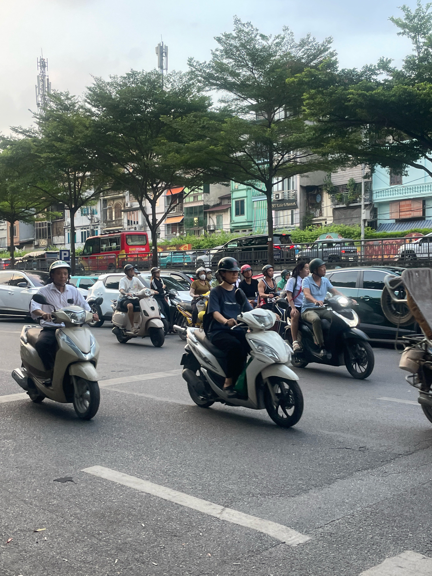The image depicts a street scene in Hanoi, Vietnam, likely during daylight hours with overcast skies. A line of motorbikes and scooters, some with riders and some with multiple passengers, are navigating a city street. The motorbikes are of various models and colors, and the riders are wearing helmets. In the background, there are buildings with a mix of architectural styles, some appearing older and others more modern. Trees line the street, providing shade. Traffic is visible, including cars and a red tour bus. The road surface is asphalt with painted white lines indicating lanes. The overall impression is a bustling urban environment typical of Hanoi.