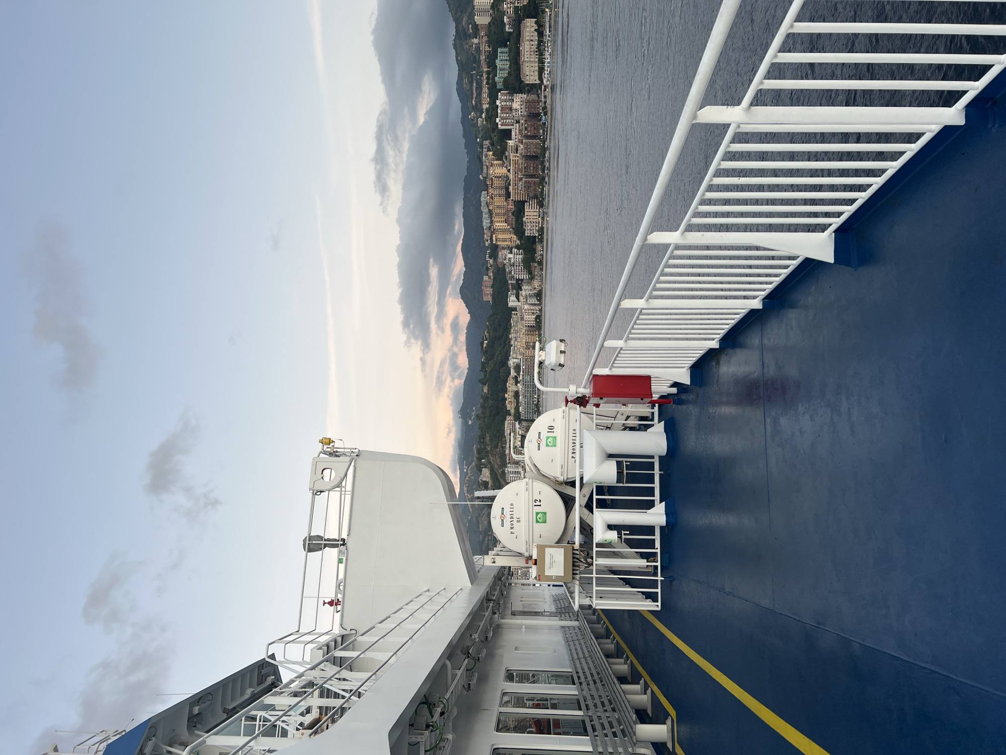 The image captures a detailed view from the deck of a ferry, with the coastal city of Messina, Italy, visible in the background. The time of day appears to be late afternoon or early evening, evidenced by the soft, diffused light and the subtle pink and orange hues on the horizon line where the sky meets the distant land.
The foreground of the image features the ferry's deck, which is predominantly dark blue with a prominent yellow safety line running longitudinally along the port side. A white metal railing with vertical balusters defines the starboard edge of the deck, overlooking the calm, dark grey-blue water. Along the port side of the deck, a series of white, slatted benches are arranged, facing outwards.
Mid-ground on the port side, two white, cylindrical life rafts are prominently displayed. Each raft bears the inscription "P MONDELLO R.C." and is numbered, with "12" on the left raft and "10" on the right. These rafts are suspended from white davits, adjacent to the ferry's white superstructure. Additional ship equipment visible in this section includes a red fire hydrant, various white ventilation pipes, and safety signage (partially obscured).
The background reveals the densely built urban landscape of Messina, situated along a coastline and extending upwards into green, tree-covered hills and mountains. Numerous multi-story buildings, primarily in shades of beige, white, and light brown, are visible, clustered along the slopes. The architectural style suggests a mix of residential and commercial structures. The sky above the city is a light blue, interspersed with scattered, wispy grey clouds.
No individuals are present or discernable within the captured scene.