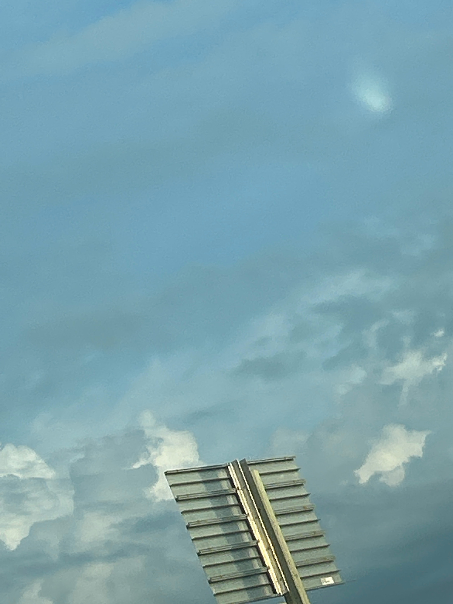 The image shows a clear blue sky with some scattered clouds. In the lower right corner, a portion of a gray, slatted sign is visible, mounted on a wooden pole. The sign is tilted upwards and to the left. The scene appears to be outdoors.