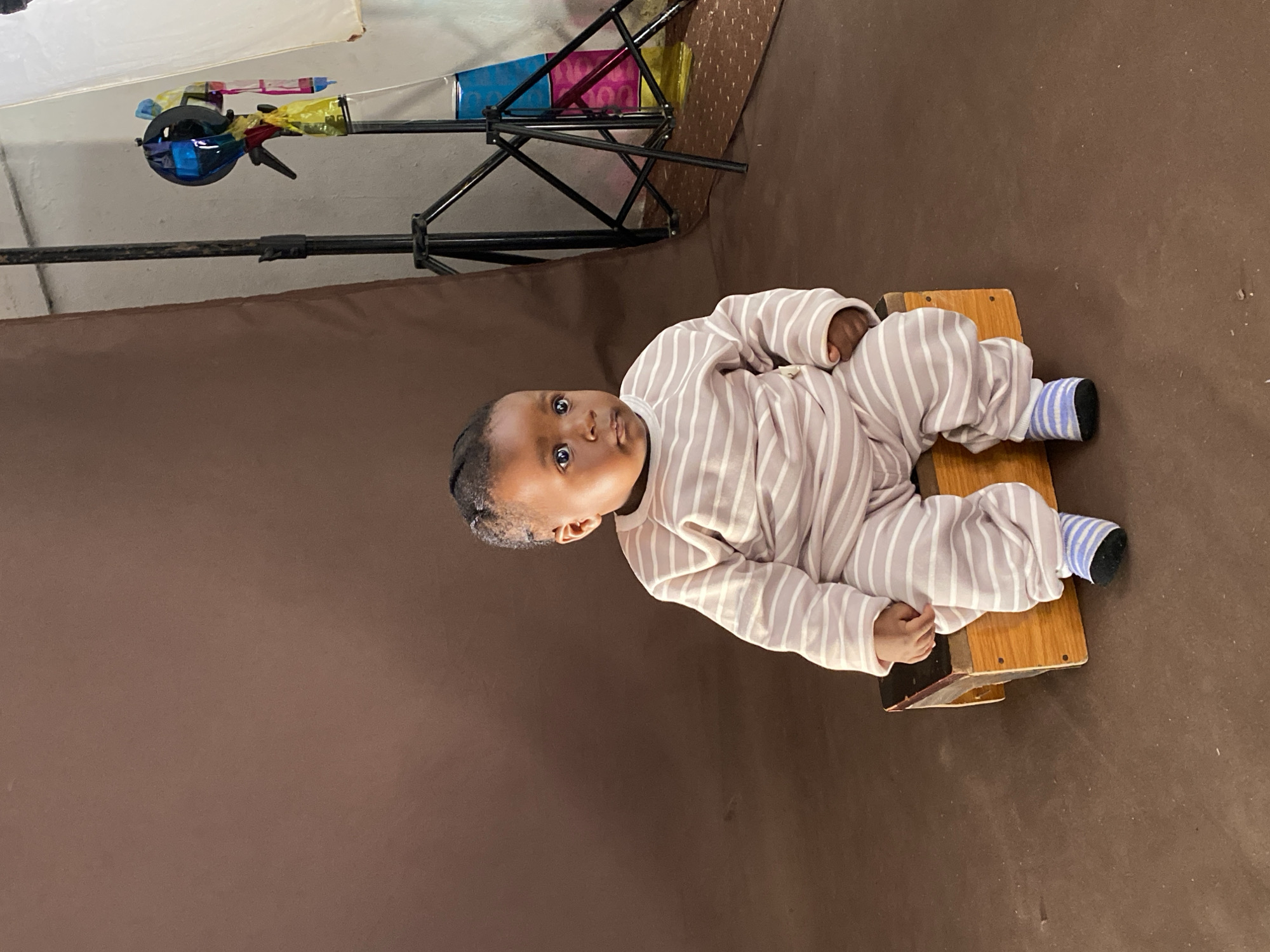 This portrait, set in Vom, Nigeria, features an infant or young toddler sitting on a small, light-brown wooden stool, positioned against a solid brown fabric backdrop. The baby, with dark skin and dark hair, is dressed in a light-colored (possibly beige or light pink) full-body striped onesie with white stripes, and blue and white striped socks. The child's body is oriented facing the camera, with its legs extended forward on the stool. The baby looks directly at the camera with wide, dark eyes and a neutral, slightly curious expression. Its hands are gently resting near its lap.
The scene suggests an indoor photography studio or a dedicated photo area. In the upper left background, behind the baby, photography equipment is visible. This includes a black light stand or tripod holding a light source, likely a flash unit, which is adorned with various colorful gels (blue, yellow, red, pink) and partially covered by a white translucent diffuser or umbrella. This setup indicates a controlled lighting environment for a professional or semi-professional portrait session. No text is visible in the image.
