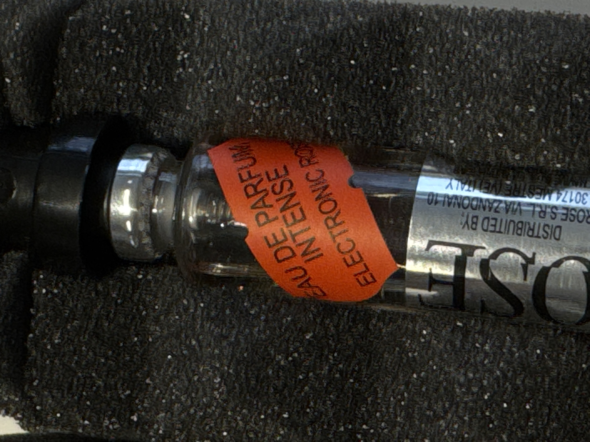 A close-up shot shows a bottle of "Electronic Rose Eau de Parfum Intense" resting on a dark, textured surface. The bottle is clear glass with a black applicator at one end. A partially obscured orange label wraps around the bottle, displaying the text "EAU DE PARFUM INTENSE ELECTRONIC RO". A silver label with the black text "OSE DISTRIBUITED BY: ROSE S.R.L. VIA ZANDONAI 10 30174 MESTRE (VE) ITALY" is visible on the lower part of the bottle. The label indicates that it is distributed by ROSE S.R.L. located at Via Zandonai 10, 30174 Mestre (VE), Italy. Mestre is a city located in the municipality of Venice, Italy, and not in Milan.