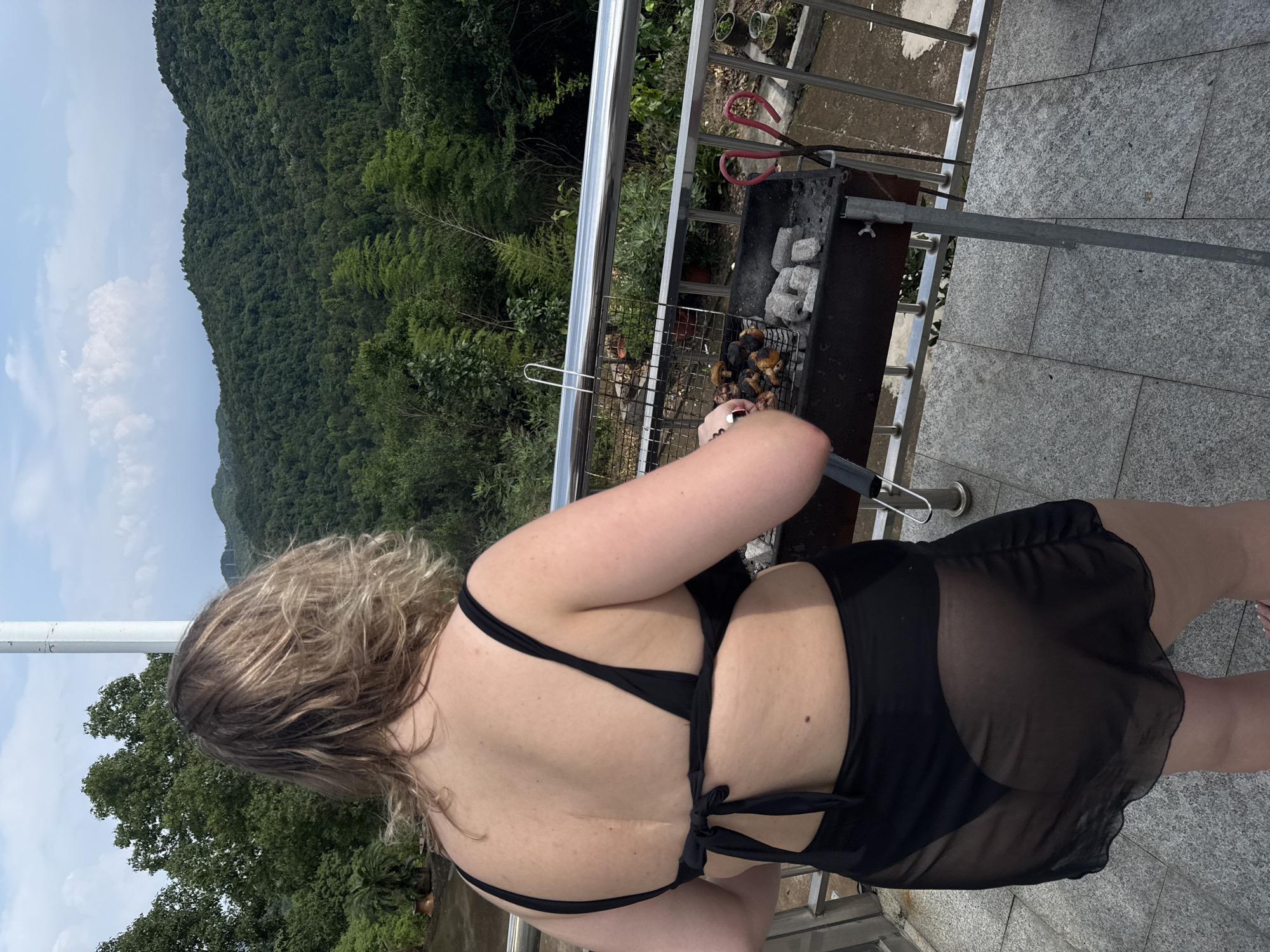 A person wearing a black bikini and a sheer black sarong is standing on a tiled balcony in front of a charcoal grill. The person is viewed from behind, and their arms are near the grill, suggesting they are cooking. The grill contains charcoal and some cooked food, possibly mushrooms and meat. There are pink-handled scissors hanging from the side of the grill. The balcony has a metal railing and overlooks a lush, green mountainous landscape under a cloudy sky. The architecture suggests this is a residential area, possibly in the city of Yiwu, China, given the context.