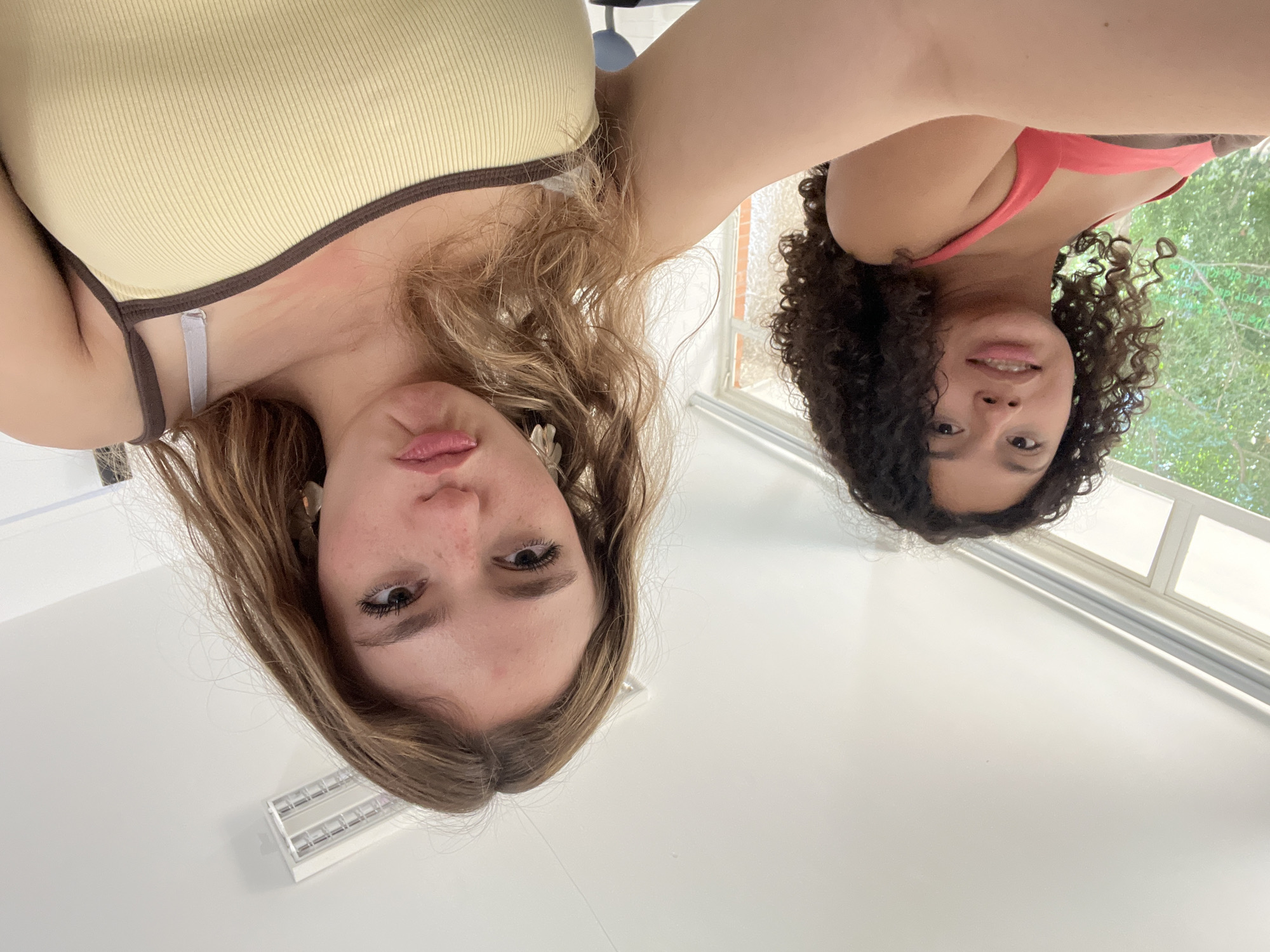 This upward-facing selfie captures two young women indoors. The image is oriented such that the subjects appear to be looking down towards the camera.
The woman in the foreground, on the left of the frame (when viewed upright), has long, wavy light brown hair and fair skin. She is wearing a light yellow ribbed tank top with brown trim, with a white bra strap visible. Her eyes are open, looking directly at the camera, and her lips are slightly pursed. A small earring is visible on her left ear.
Behind her, on the right side of the frame, is a second woman with dark, curly hair and darker skin. She is wearing a coral or pink sports bra or tank top. She also looks towards the camera with open eyes and a slight smile, revealing her teeth.
The background primarily consists of a white ceiling and walls. A rectangular light fixture is visible on the ceiling above the first woman. To the right, a window allows natural light into the room, showing lush green foliage outside, suggesting a daytime setting.
The overall impression is a casual, friendly selfie shared between the two individuals. There is no visible text in the image.