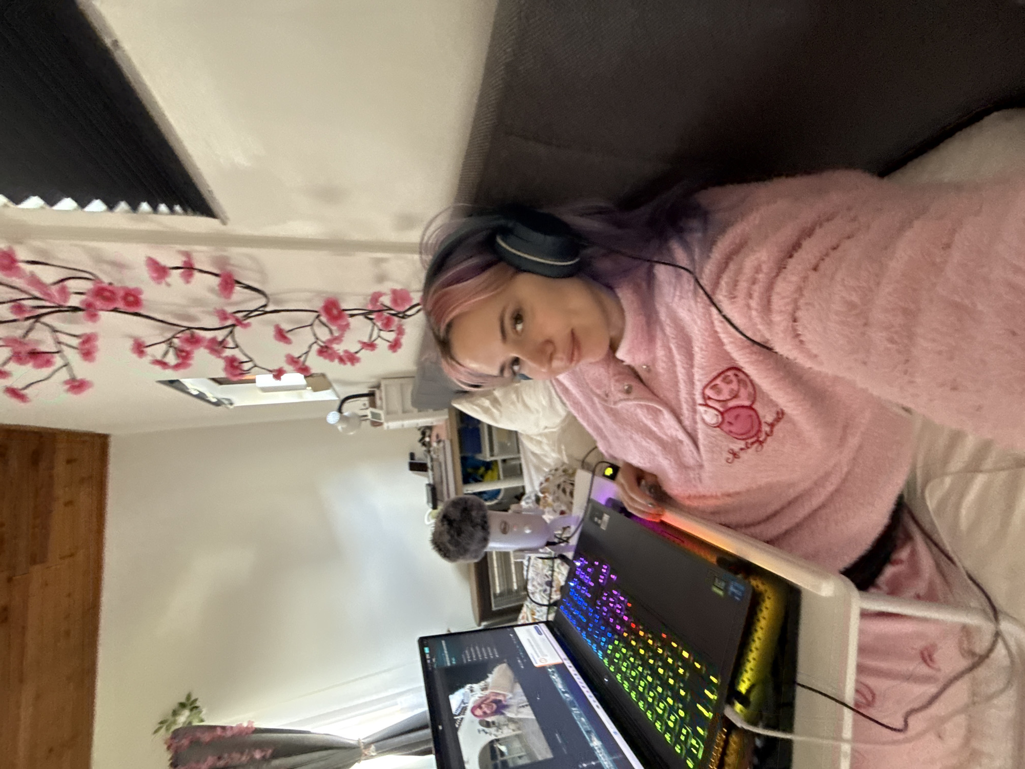 A female subject is depicted indoors in the City of Zagreb, Croatia, seated on a gray sofa or bed, leaning against the backrest. She has light skin, multi-colored hair (pink and purple hues), and wears a pink fluffy long-sleeved top with a cartoon-style embroidered motif on the chest. Dark blue over-ear headphones are worn over her ears. She is looking directly towards the camera with a slight smile.
In front of her, a black laptop with an illuminated RGB keyboard rests on a white, portable bed desk. The laptop screen displays a video editing interface featuring a video clip of a female subject with pink hair in an outdoor setting. To the right of the laptop, a light-colored Blue Yeti USB microphone with a gray fluffy windscreen is positioned on the desk.
The room features white walls, with a sloped ceiling of exposed light-colored wooden planks. On the left, a window with light-colored curtains, possibly gray, is partially visible, tied back, with a small artificial green plant near the top. On the right, a white wall is adorned with a vertical decorative element resembling a branch with numerous pink artificial flowers. Further right, a white shelving unit with a white desk lamp is present, and a dark roller blind is partially visible at the upper right. Multiple black and white wires are visible on the bed and desk.
