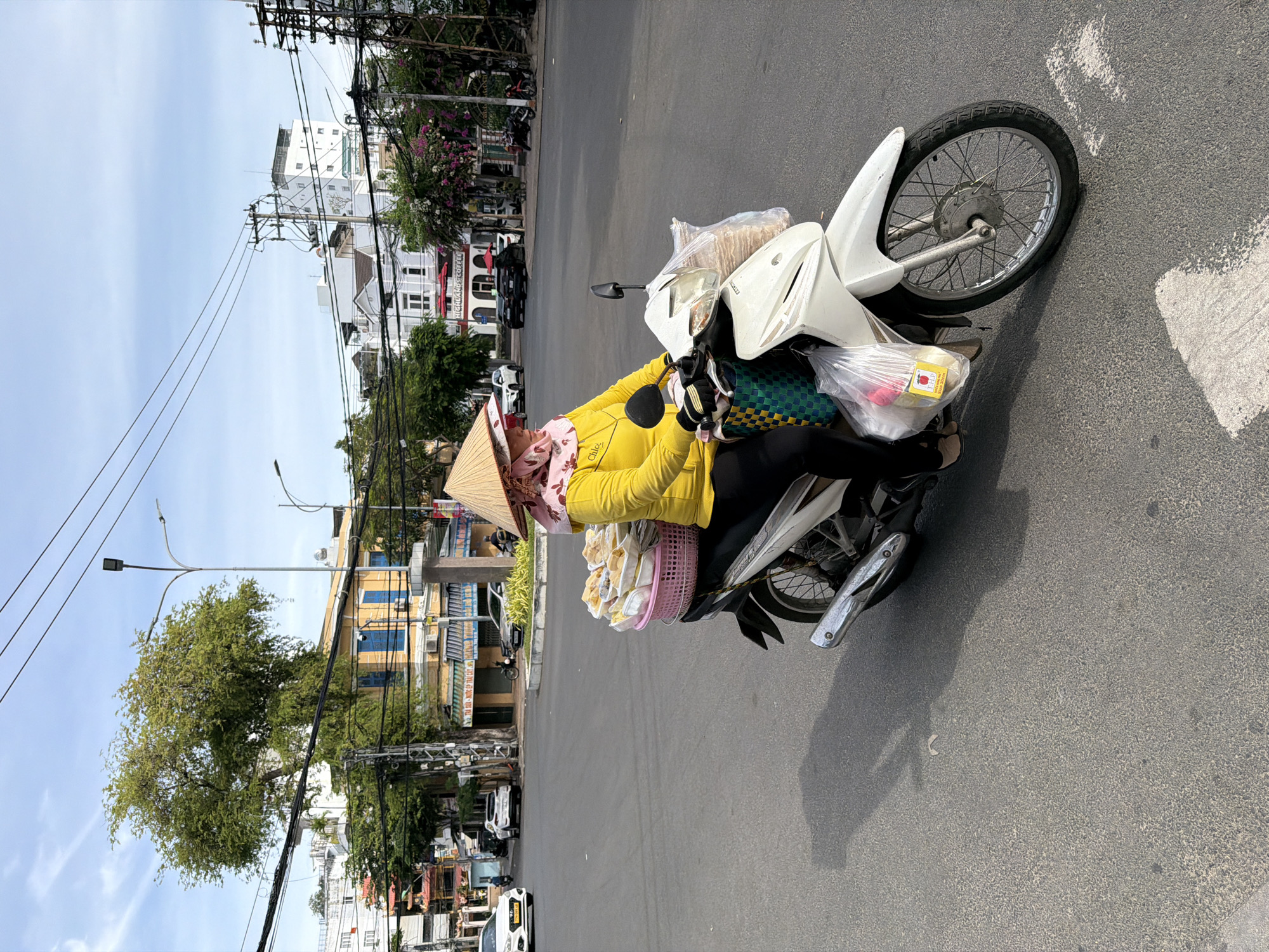 A woman wearing a yellow long-sleeved shirt, black pants, and a conical hat rides a white motorcycle on a street in Nha Trang, Vietnam. She is carrying a pink basket filled with food items on the back of her motorcycle. The street is paved and appears to be an intersection, with buildings and trees visible in the background. There are power lines overhead and several cars and other motorcycles on the road. The sky is partly cloudy.