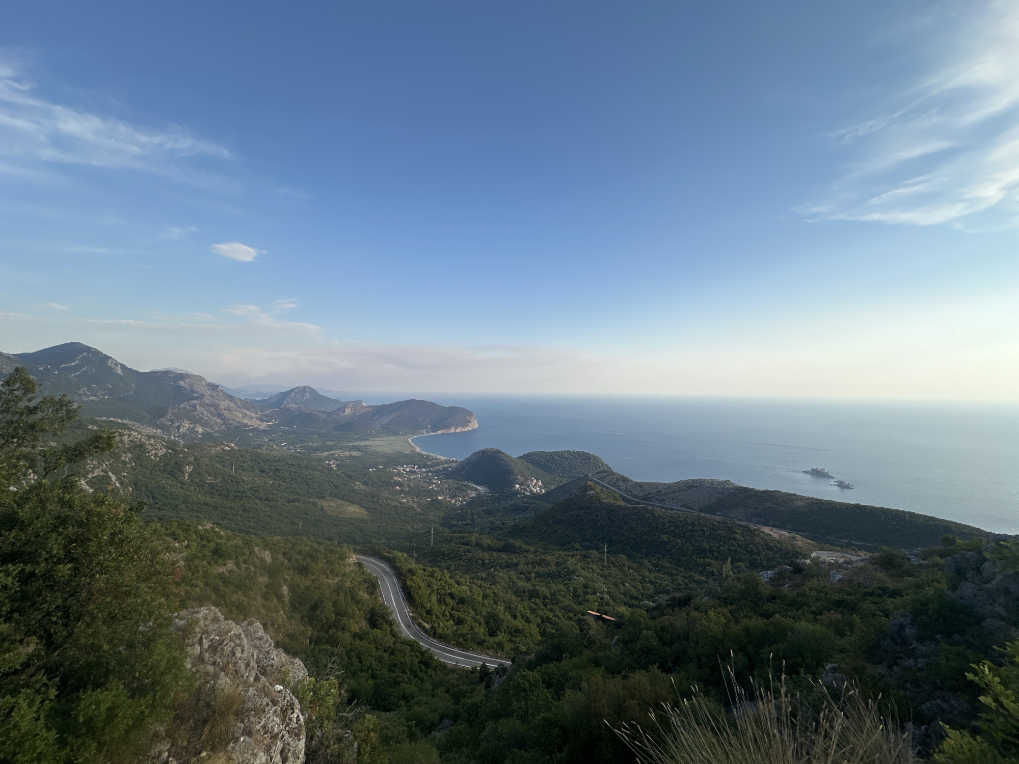 The image depicts a scenic overview of the coastline and surrounding mountainous terrain near Novoselje, Montenegro. A winding asphalt road carves its way through lush green hills, disappearing into the dense foliage. In the distance, the azure sea stretches out to the horizon, dotted with a few small islands. Several buildings are visible nestled within the valleys and along the coastline, suggesting a small settlement. The sky is a clear blue, with a few wispy clouds scattered across it, indicating fair weather. The overall scene is one of natural beauty and tranquility.