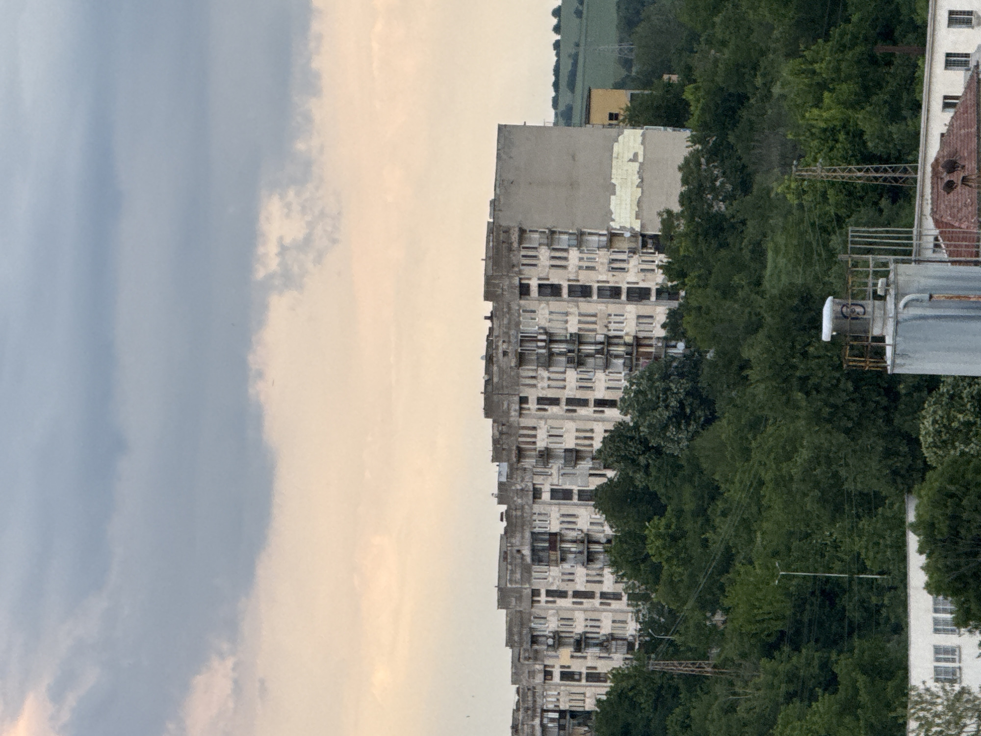 The image shows a high-angle view of an apartment building in Lovech, Bulgaria. The building is light-colored with multiple stories and rows of windows. The building has some damage on top of it. A row of green trees separates the apartment building from a white building on the right. The sky in the background is partly cloudy.