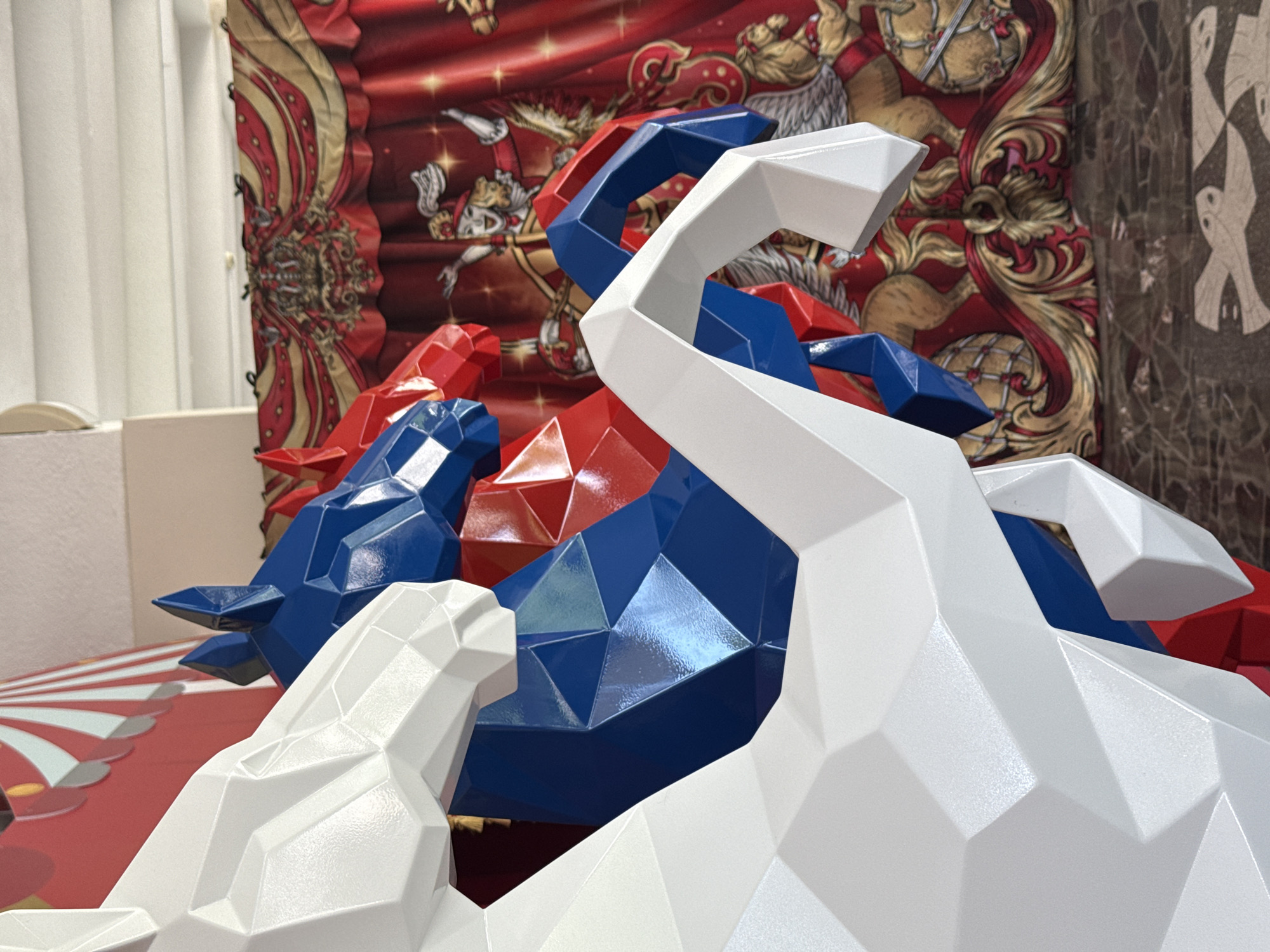 The image captures a close-up view of three abstract horse sculptures rendered in white, blue, and red, positioned in front of a vibrant circus-themed tapestry. The horse sculptures, characterized by their sharp, geometric facets, are layered in the foreground, with a white sculpture dominating the left side, partially obscuring a blue and red sculpture. The tapestry in the background features a detailed scene with a ringmaster figure, horses, and ornate red and gold patterns, evoking a circus atmosphere. The overall setting appears to be indoors, possibly a venue or exhibition space. The floor is a dark, patterned mosaic. The lighting suggests an interior environment with diffused light from above. The specific location in the city of Sochi, Russia, is not identifiable from the visual information presented in the image.