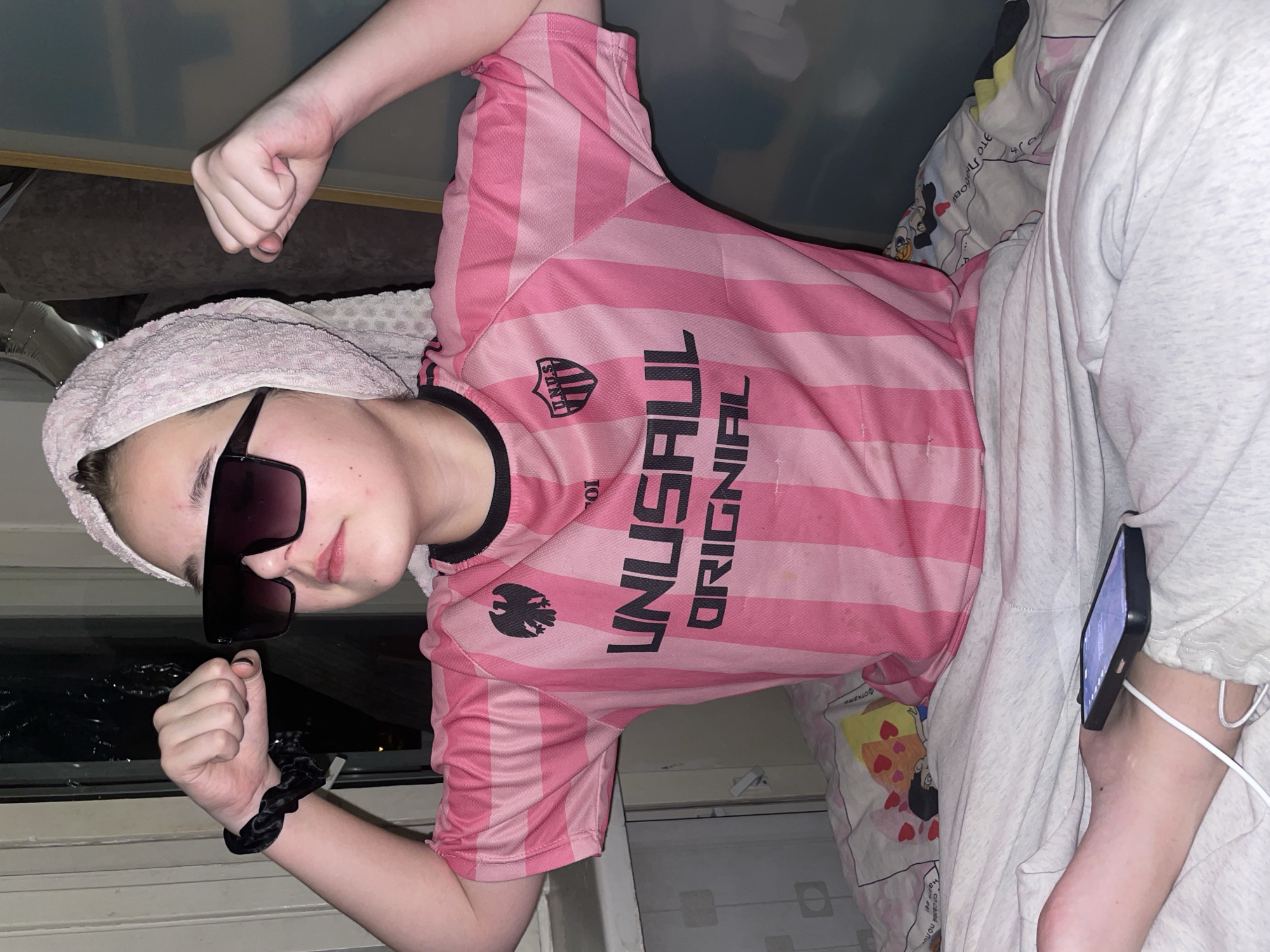 A young person is captured in a playful indoor portrait, lying down on what appears to be a bed or sofa. The image is oriented with the subject rotated 90 degrees clockwise.
The subject is wearing a pink and white striped short-sleeved t-shirt with black trim, and light grey sweatpants. A light pink terry cloth towel is wrapped around their head, and oversized dark sunglasses obscure their eyes. Both hands are clenched into fists and held up, suggesting a humorous or confident pose. A black scrunchie is visible on their left wrist.
In the background, colorful pillows with various patterns, including cartoonish designs and one with yellow, red, and heart motifs, are visible. A dark window or reflective surface to the lower left suggests it is nighttime or very dim outside. The room itself is dimly lit. A black smartphone, connected to a white charging cable, is propped on the person's right ankle, indicating they might be viewing content while relaxing.
Visible text on the pink shirt includes "UNUSUAL ORIGINAL" and a logo containing "UNUSUL" within a shield shape, all in black capital letters. Partially visible Cyrillic text like "ОТО" and "БОЛЬ" can be seen on one of the pillows. The scene conveys a relaxed and lighthearted mood. The location context is Vsevolozhsk, Russia.