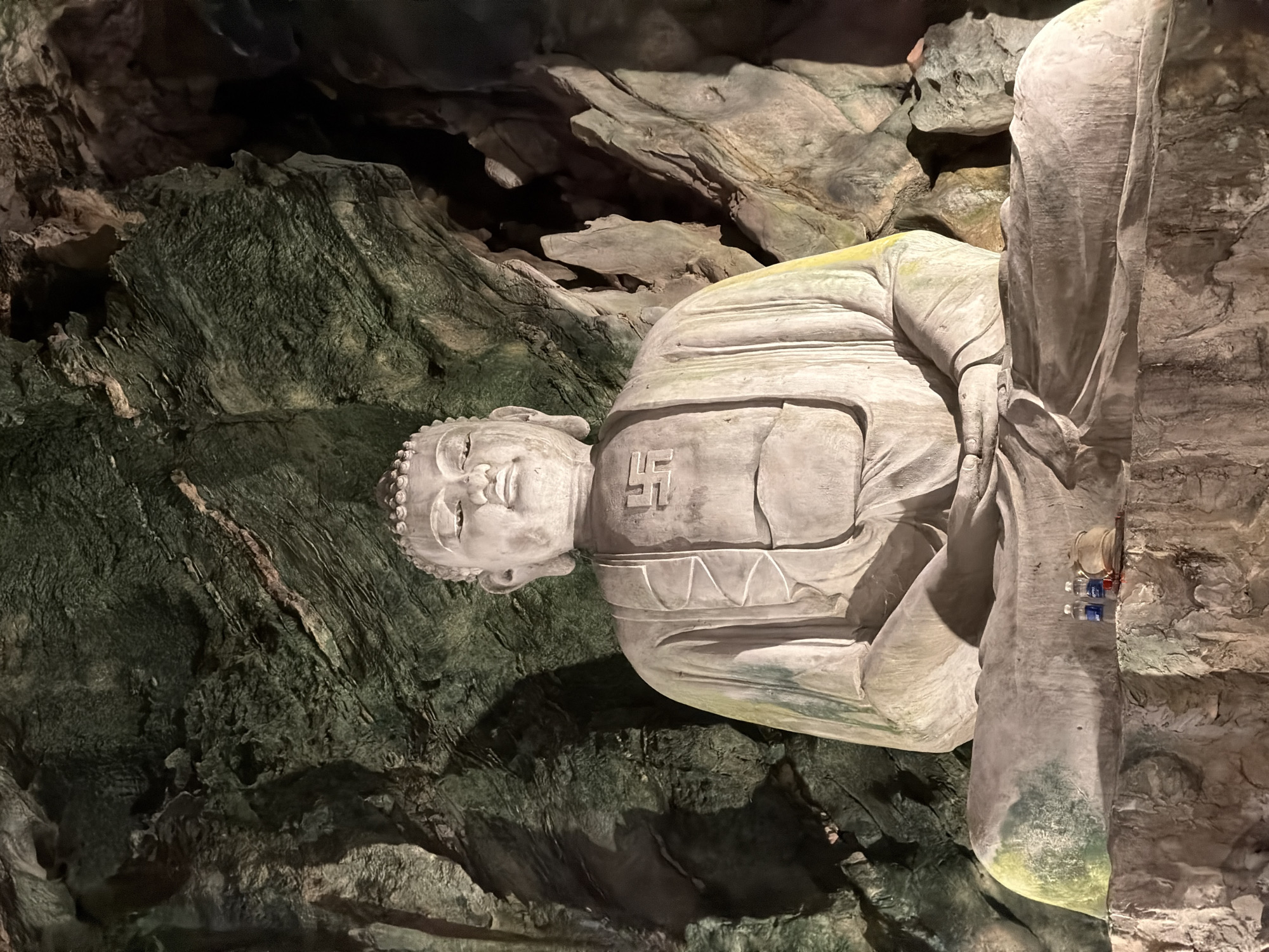 The image shows a reclining Buddha statue within a grotto or cave-like setting. The statue is light in color, possibly made of stone or concrete, and is positioned with its head oriented to the left. Distinctly, a swastika is visible on the chest area of the statue. The statue is set against a backdrop of rough, uneven cave walls that are varied in color, ranging from dark green to beige. The right side of the statue is adjacent to a rocky wall with some small items affixed to it, possibly bottles or religious offerings. This scene is found in one of the cave temples of the Marble Mountains in Đà Nẵng, Vietnam.