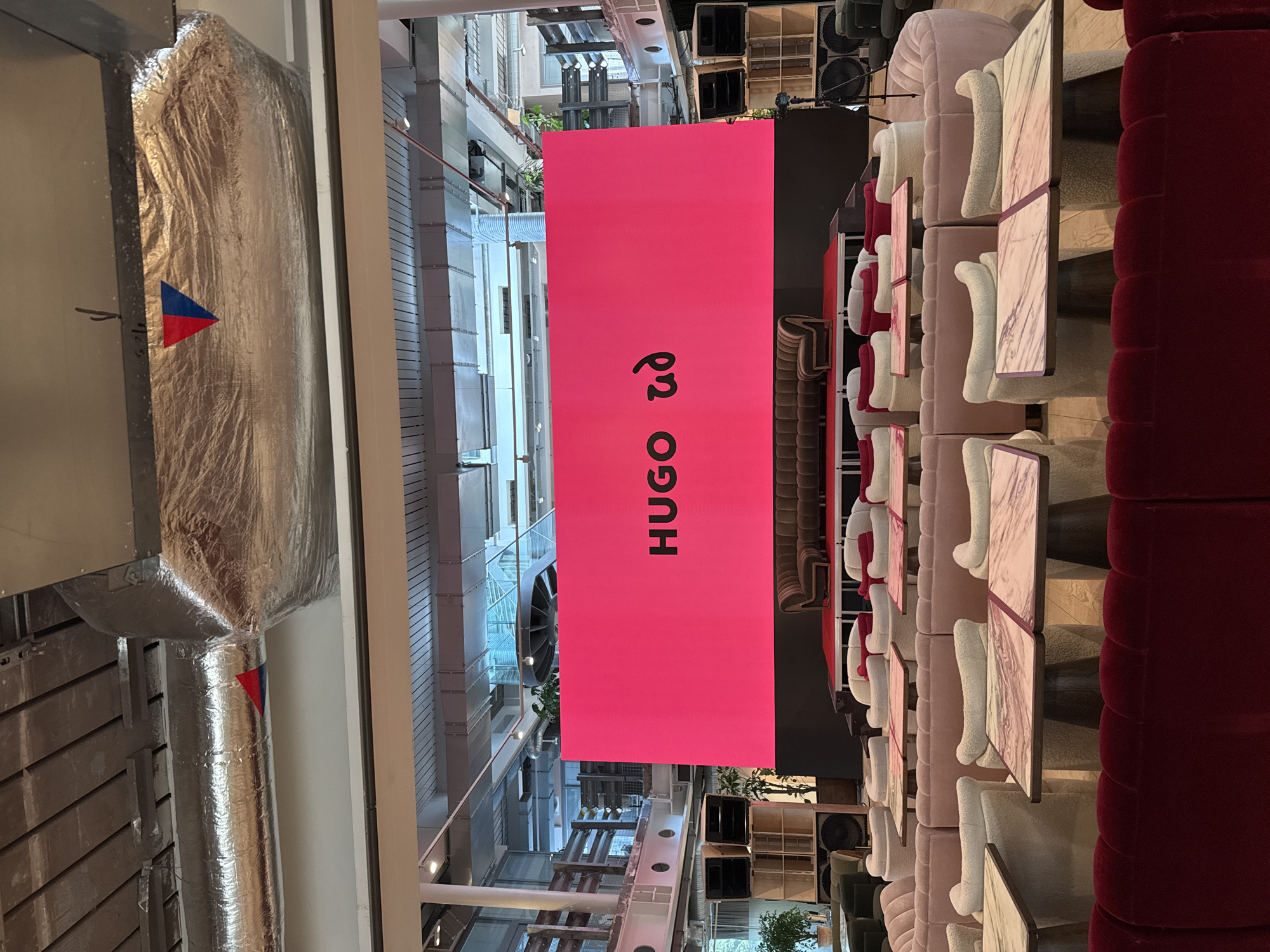 The image depicts the interior of a commercial space, possibly a restaurant or café in London, United Kingdom. A prominent pink banner displaying the word "HUGO" in black lettering, partially written in an oriental script, dominates the view. The banner hangs in a multi-story area with exposed architectural elements and utility systems. To the right, a line of tables with chairs and plush seating in varying shades of pink and red creates a dining or lounge area. The space exhibits a modern design with a blend of industrial and upscale aesthetics. There are no people visible in the shot. The architectural context suggests a commercial or retail environment in a city center.