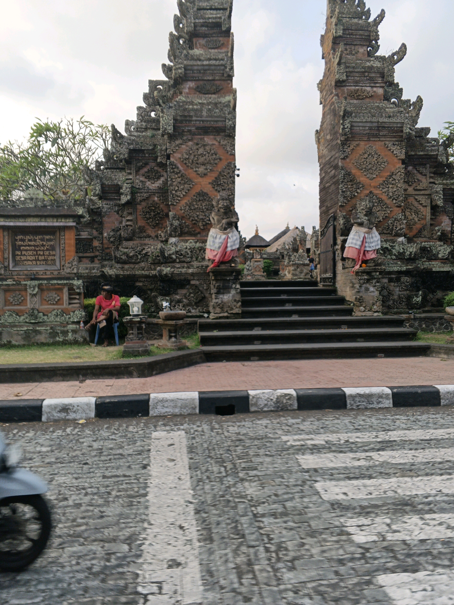 This is a full-shot, eye-level, outdoor, daytime photograph of the Pura Puseh Desa Batuan temple entrance in Sukawati, Indonesia. The temple features ornate, weathered stone architecture with intricate carvings. Two towering gateway structures flank a wide set of stairs. A man in a bright red shirt sits on a blue plastic chair to the left of the stairs, facing away from the camera. He is holding an object in his lap. In the foreground, a textured cobblestone street with a pedestrian crossing is visible, with the blurred front wheel of a motorcycle on the left side of the frame. The sky is overcast.
