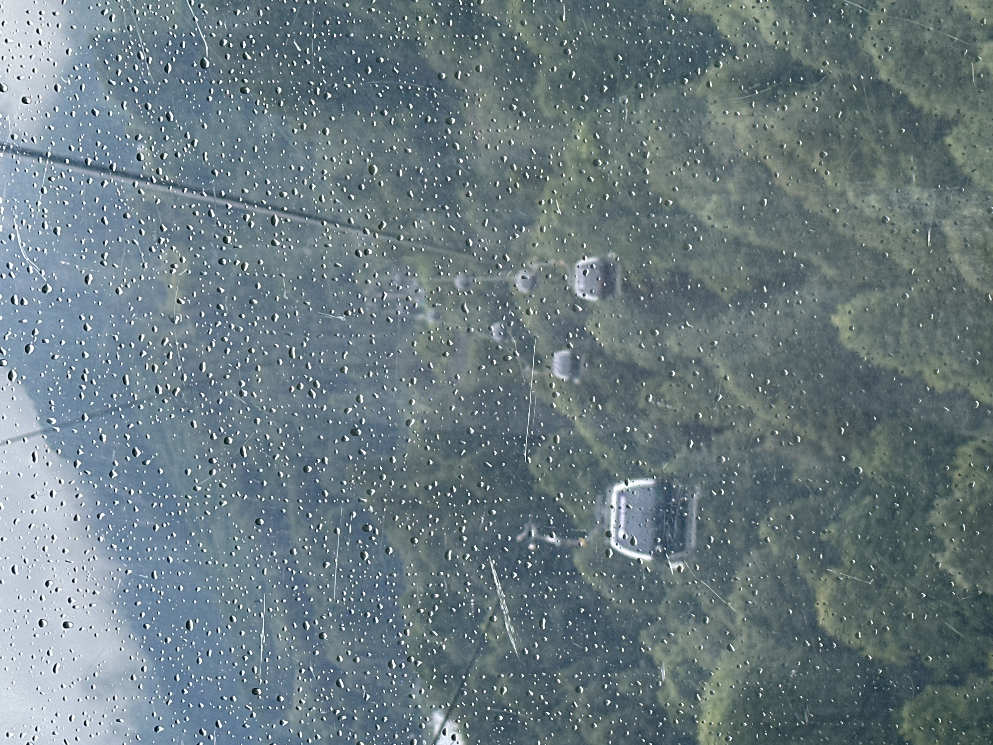 The image, taken in Estosadok, Russia, shows a view through a rain-streaked window of a mountain landscape. Several cable cars are visible, suspended on wires above a dense forest. The cable cars, appearing as grey, rounded cabins, are spaced out along the cables, suggesting they are in transit. The forest below is a uniform green, indicating a thick canopy of trees covering rolling hills or mountains. The sky appears overcast, contributing to the muted color palette of the scene. The rain droplets on the window create a textured, blurred effect, obscuring some of the finer details of the landscape and the cable cars. The overall impression is one of a moody, wet day in a mountainous region.
