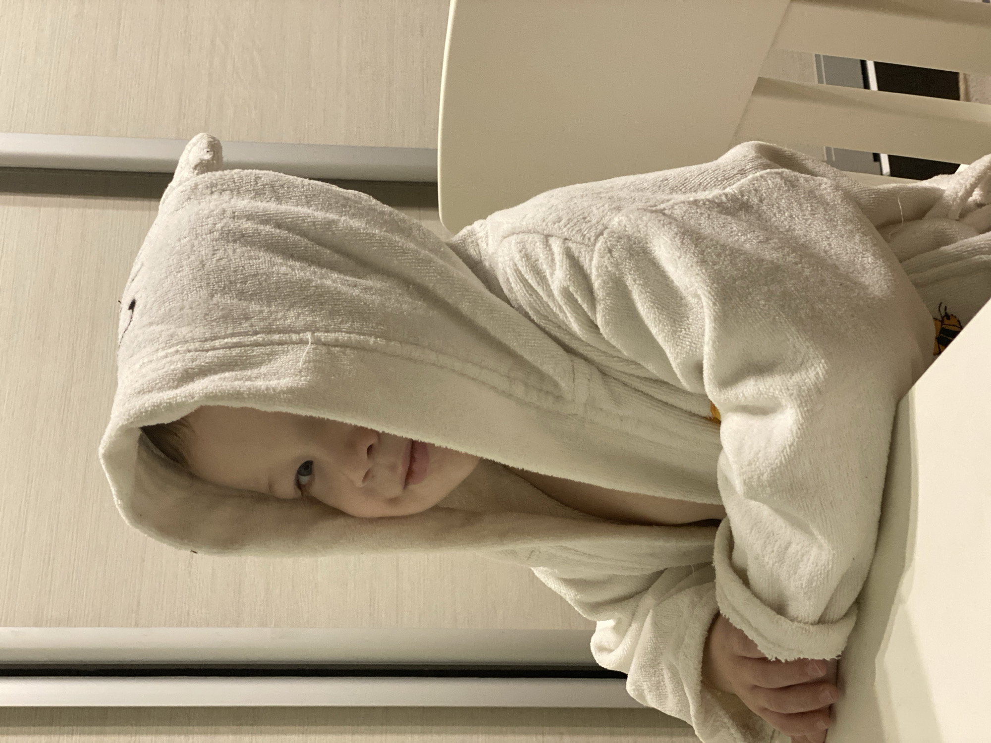 A young child wearing a white hooded bathrobe is partially visible, peering out from under the hood. The child appears to be positioned in or near a white structure, possibly a crib or bed frame. The background includes a light-colored wall with a horizontal stripe. The image does not provide any information on whether the picture was taken in Sochi, Russia. There is no information regarding people or subjects involved, actions, interactions, or the environment.