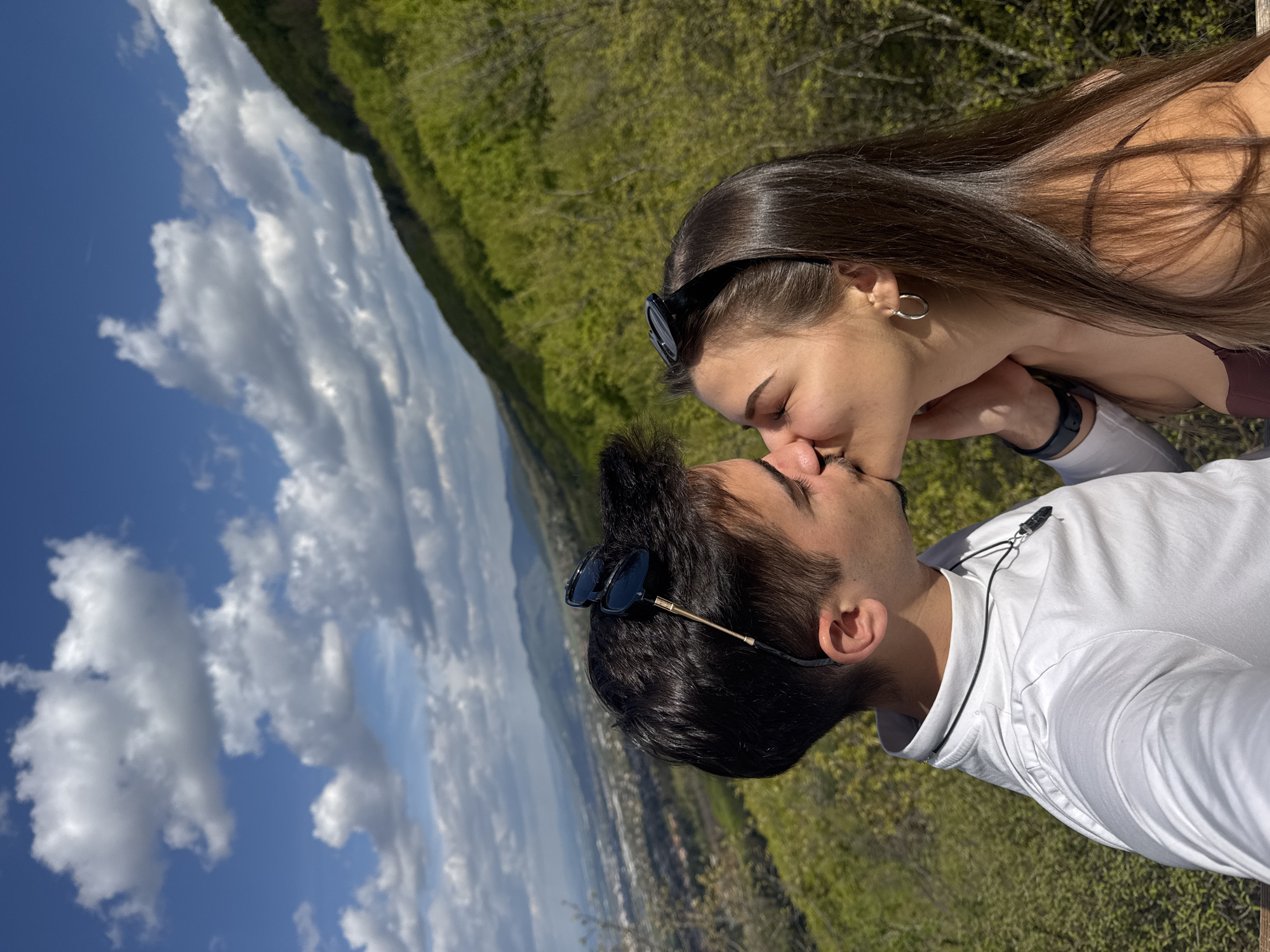 A couple is kissing on a scenic overlook. The woman has long, dark hair and is wearing sunglasses on her head, a hoop earring, and a dark-colored top. The man has dark hair, sunglasses on his head, and a white shirt with a black lanyard around his neck. They are positioned in front of a green, hilly landscape and a blue sky with white clouds. In the background, a cityscape can be seen. The image was taken at Boyana Lake, Bulgaria, a suburb of Sofia.