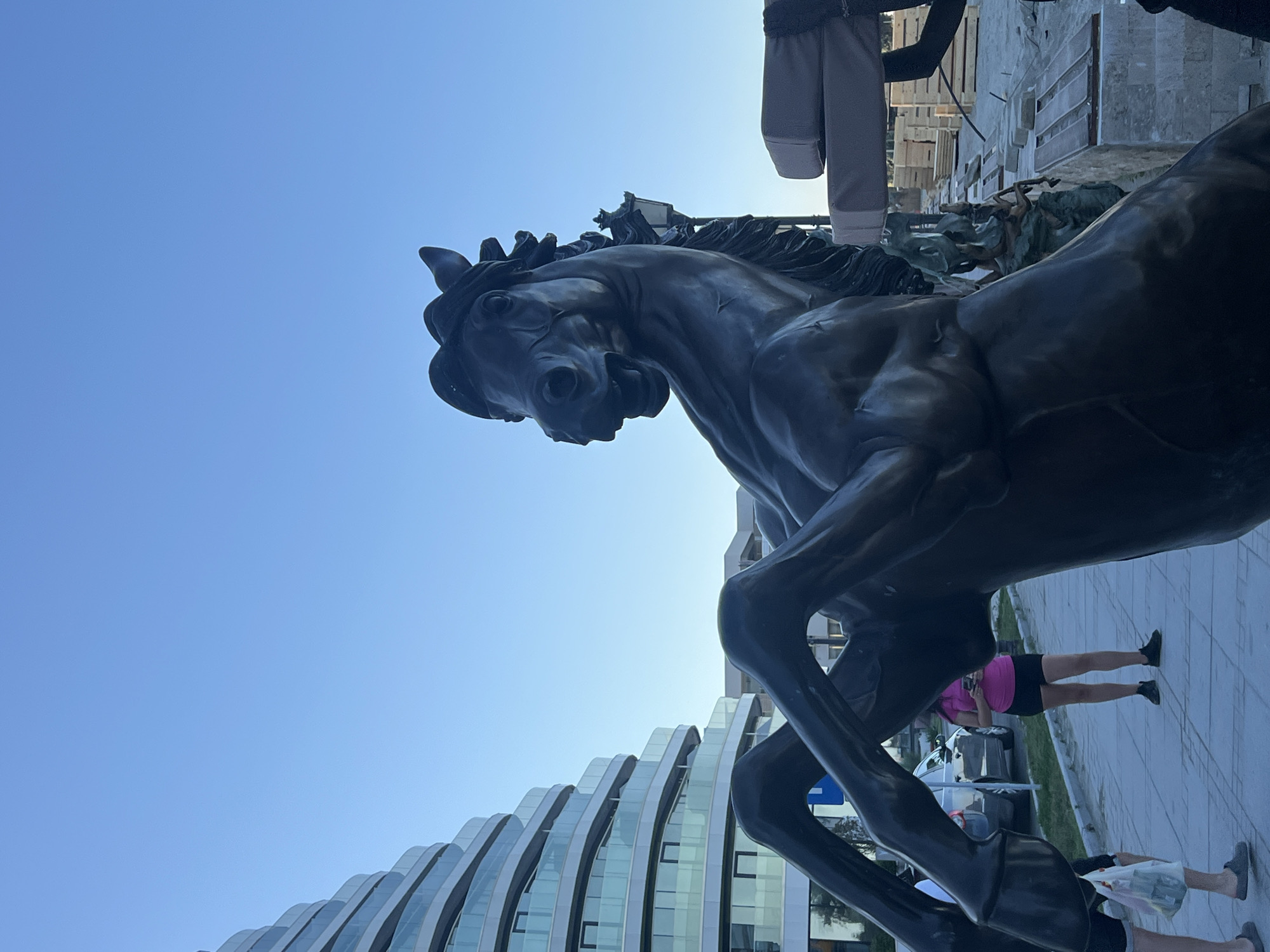 This is a photograph taken in Năvodari, Romania, showing a large bronze statue of a rearing horse in the foreground. The horse is depicted with its head turned to the left and its mouth open. Behind the statue, a modern, curved building with multiple balconies and glass railings is visible on the left side of the image. To the right of the statue, there is a street lamp and some construction materials, including stacks of wooden planks and what appear to be benches. In the middle ground, a woman in a pink shirt and black shorts is standing on a paved walkway, looking at her phone. Other individuals are partially visible in the lower left corner of the image. The sky is clear and blue, suggesting it is daytime, possibly late afternoon or early evening due to the lighting. The overall scene appears to be an outdoor public space, possibly a promenade or plaza near the coast.