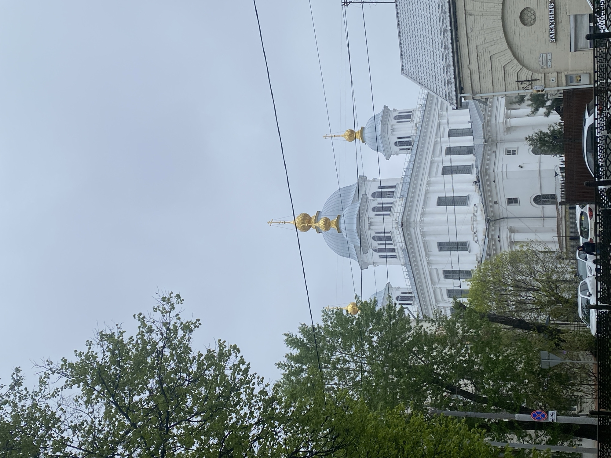 The image shows a partial view of the Holy Cross Cathedral in Заволжье, Russia. The cathedral is white with blue domes topped with golden crosses. The sky is overcast. Power lines are visible in the foreground. A brick building stands to the right of the cathedral. Trees with green foliage are in the foreground. Cars are parked along the street, and a person is walking near them. A fence is also visible along the street.