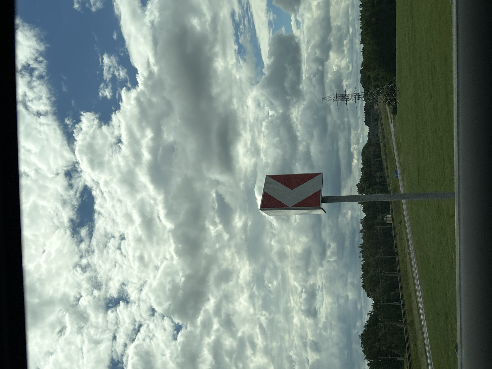 The image depicts a roadside scene under a cloudy sky. In the foreground, a grey pole supports a traffic sign indicating a sharp left turn, with alternating red and white chevron patterns. To the left of the sign, a railway track runs parallel to a road. A dense line of green trees forms the horizon, with a tall electricity pylon visible in the distance on the right. The terrain is a grassy field, suggesting an outdoor, possibly rural, setting. The overall lighting suggests it is daytime. The scene appears to be in the vicinity of Малевіцкі сельскі Савет, Belarus, given the prompt's context.