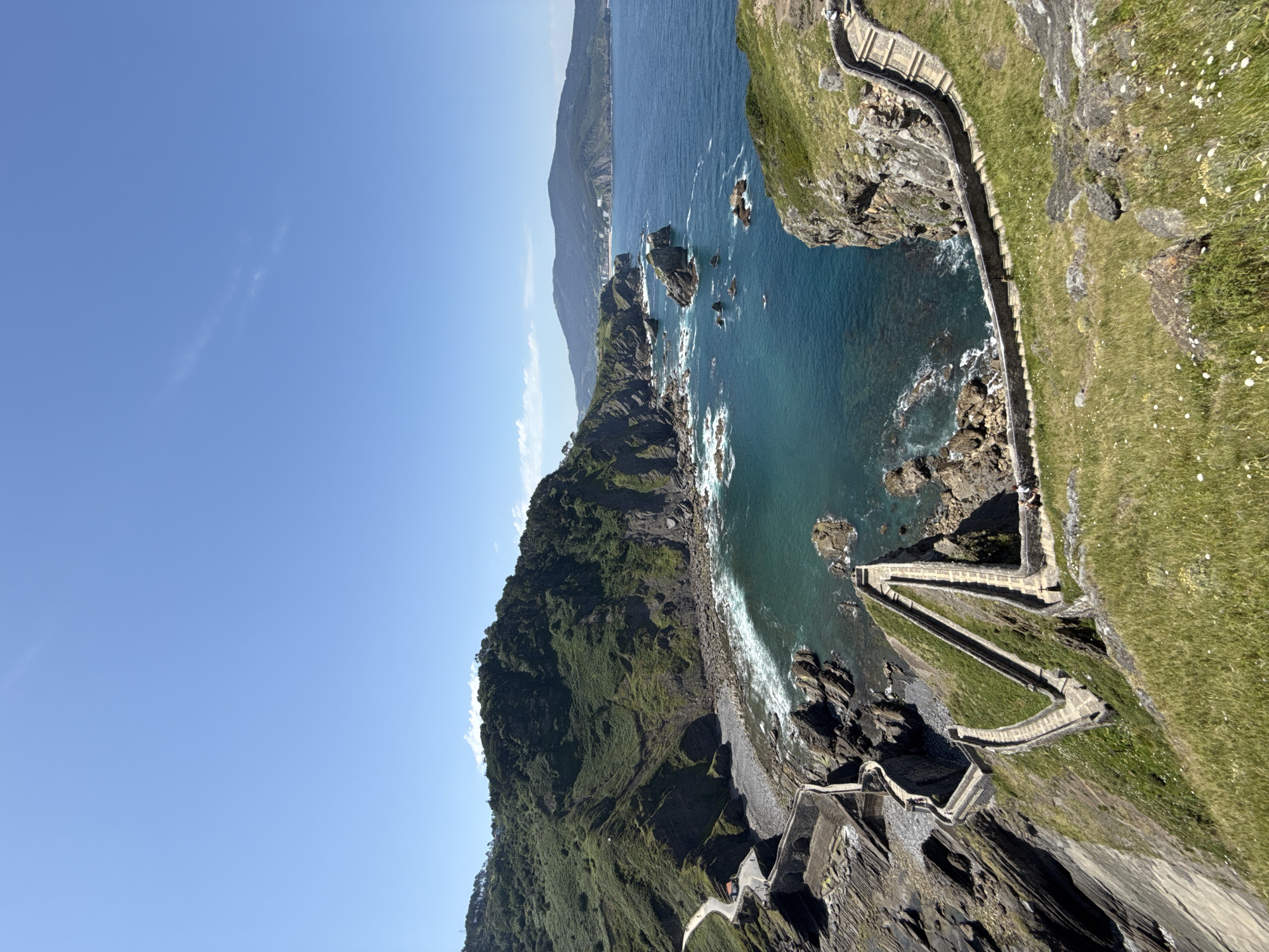 The image shows a scenic view of the coast of Bakio, Spain, featuring the iconic San Juan de Gaztelugatxe steps. The steps, made of stone and winding in a zig-zag pattern, ascend a steep, grassy hill that leads to the sea. The turquoise water of the ocean crashes against rocky shores and sea stacks. Lush green vegetation covers the surrounding hillsides. In the distance, more hills and a town can be seen along the coastline under a clear blue sky. There are a few people visible on the steps and the grassy areas, observing the landscape. The overall scene captures the dramatic beauty of the Basque coast.