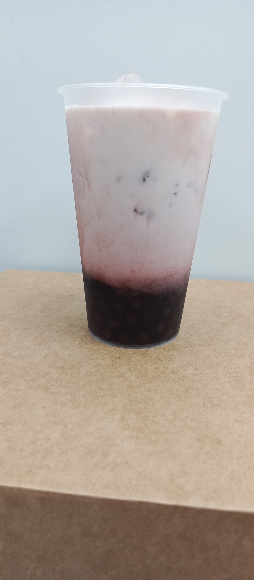 A clear plastic cup filled with a layered beverage sits on a brown surface. The beverage has a dark, grainy layer at the bottom, a reddish-pink transition layer in the middle, and a pale pink, milky layer at the top. There are small, dark specks visible throughout the milky layer, and a piece of ice pokes out from the top. The background is a plain, light blue wall. The scene is captured from a slightly low angle, focusing on the cup and its contents. The location in Bugry, Russia is not discernible from the image.