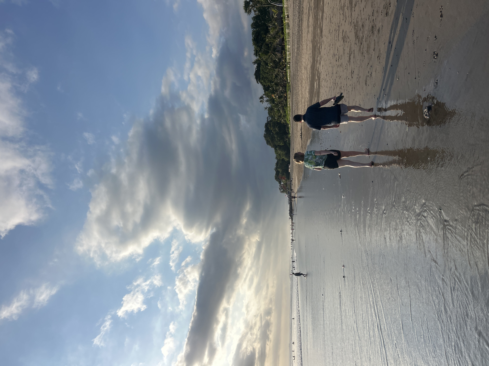 The image depicts a wide, sandy beach at Kuta, Indonesia, during a time of day that suggests either late afternoon or early morning, given the low angle of the sun and the dramatic cloud formations in the sky. The sky is a prominent feature, filled with large, textured clouds, some illuminated by sunlight, creating a contrast between bright and shadowed areas. The sun is positioned to the left of the frame, casting a warm glow.
In the foreground, two individuals are walking away from the camera along the wet sand near the waterline. The person on the left is wearing a green patterned short-sleeved shirt and black shorts, while the person on the right is in a dark short-sleeved shirt and gray shorts, carrying a pair of dark sandals in their right hand. Their reflections are visible in the shallow water covering the sand.
Further down the beach, numerous other people are scattered along the shore and in the water. Some are walking, while others appear to be standing or engaged in activities near the gentle waves. The distant shoreline is lined with lush green vegetation, including palm trees, and hints of buildings or structures can be seen nestled amongst the trees. The wet sand reflects the light, creating a smooth, shimmering surface. The overall scene conveys a sense of calm and leisure.