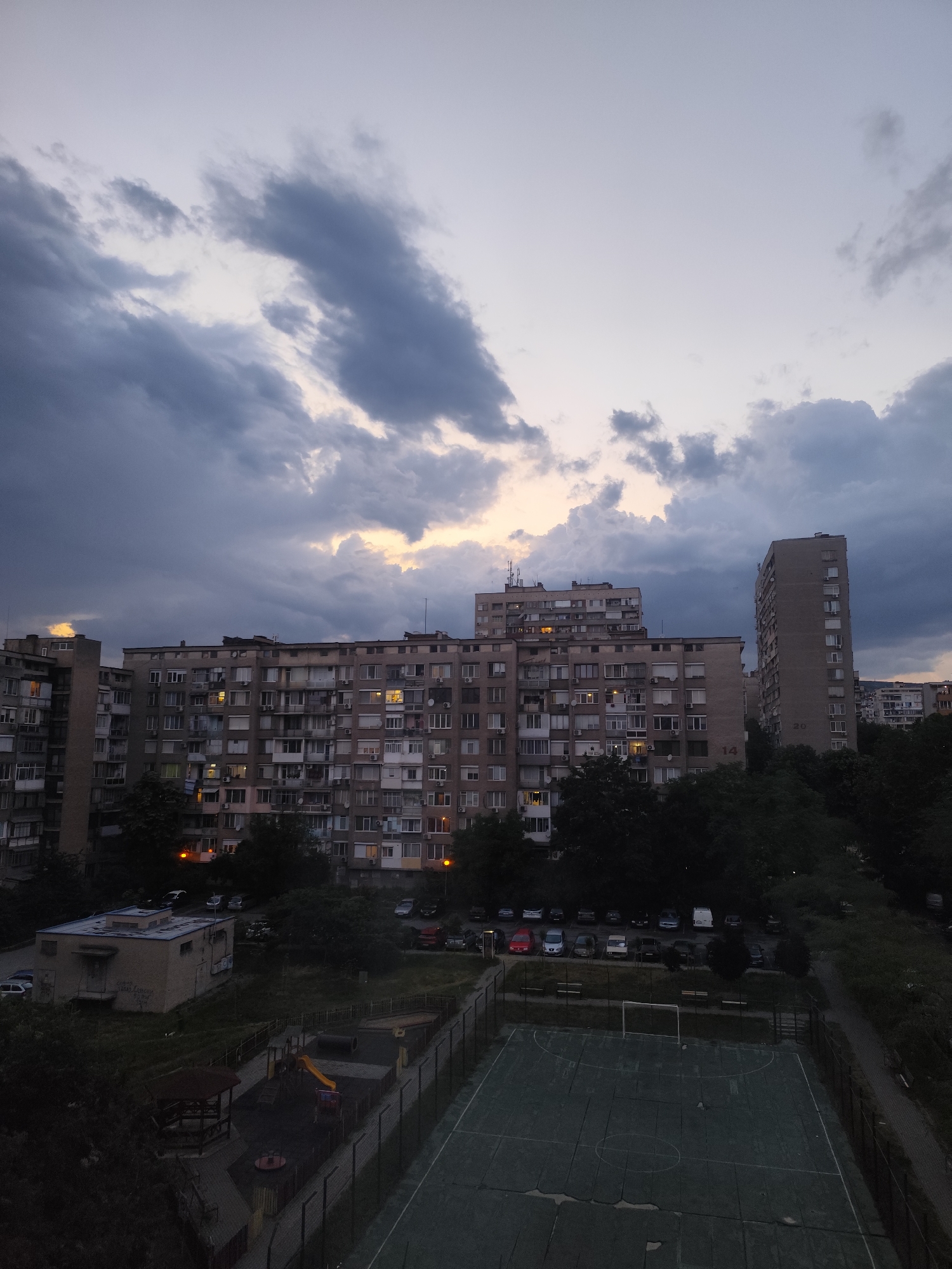 The image depicts a residential area at dusk, likely in Stara Zagora, Bulgaria, based on the architectural style of the buildings. A series of large, Soviet-era apartment buildings dominate the midground. Some windows are illuminated, suggesting occupancy. A parking lot with multiple vehicles is visible in front of the buildings. Below, there is a playground and a sports field, both seemingly empty. The sky is overcast with dark, heavy clouds, and a faint golden light peeks through, possibly from the setting sun. The scene appears calm and quiet, with no visible human activity.