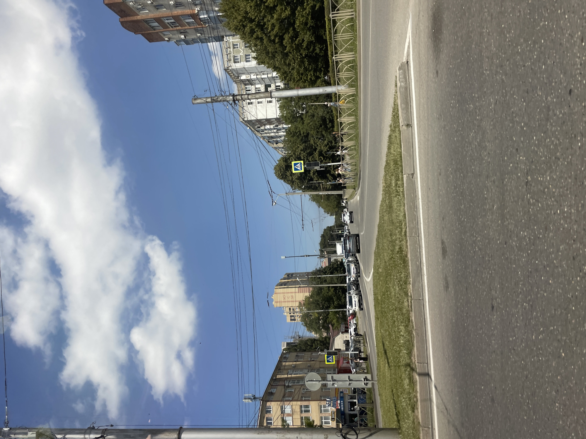 The image depicts a city street scene in Stavropol, Russia, on a clear day. In the foreground, a paved road with white lane markings is visible, leading into the mid-ground where several cars are driving. On the left side of the road, a sidewalk area features a pedestrian crossing sign with a blue background and a yellow pedestrian symbol, alongside a lamppost and trees. Adjacent to the sidewalk is a grassy area with a curb.
Further into the mid-ground and background, multi-story apartment buildings line the street. These buildings exhibit typical Soviet-era architecture, with some constructed from brick and others from concrete panels. Trees are interspersed between the buildings and along the roadside, indicating a developed urban environment. Overhead, a network of utility wires is strung between poles, a common sight in Russian cities. The sky is a vibrant blue with scattered white clouds.
People are visible on the sidewalks, appearing to be pedestrians walking or waiting. The overall impression is a busy urban street with ongoing traffic and daily life unfolding under a pleasant sky.