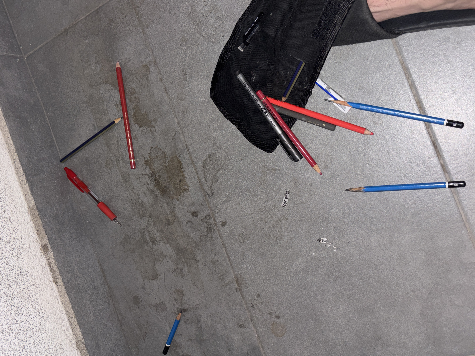 The image shows a collection of pencils, pens, and a black pouch or bag on a grey tiled floor. The objects are scattered around the bag, suggesting they may have fallen out. A red pen, a blue pencil, a red pencil, and a dark-colored pencil form a rough triangle on the left side of the image. Several pencils of varying colors and a mechanical pencil are clustered near the bag. One blue pencil is isolated near the bottom of the image. The setting is a tiled floor, possibly indoors. The location is uncertain, but not defined within the current image. There are no people.