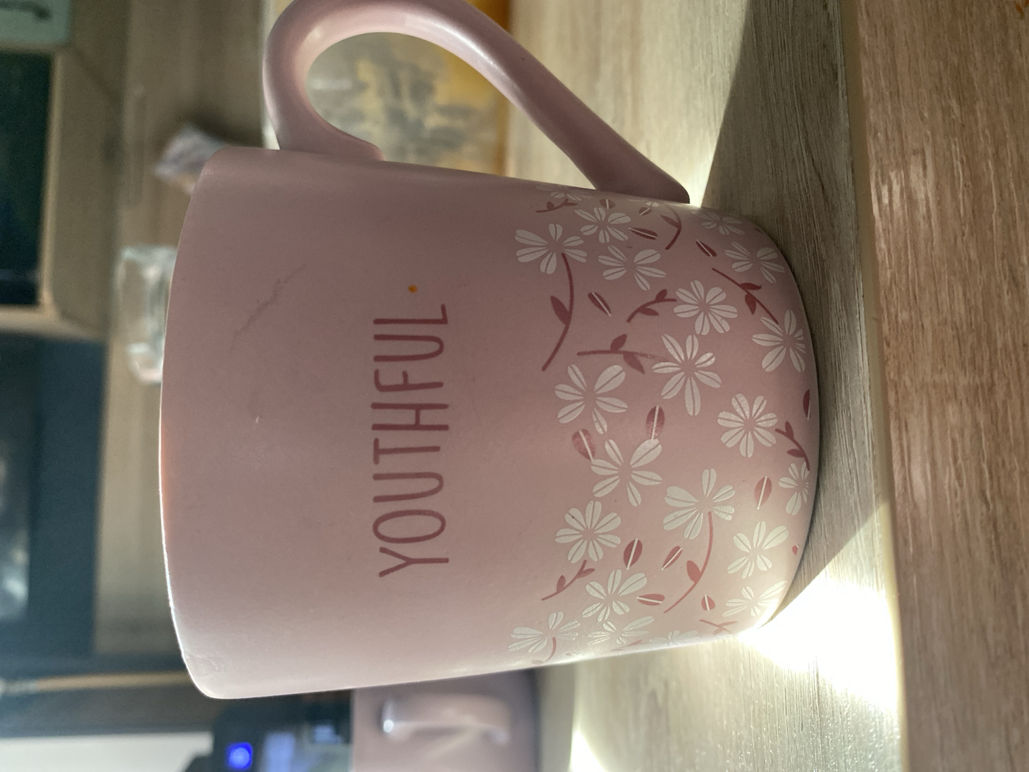 A pink mug with the word "YOUTHFUL" printed vertically on it and decorated with white flowers and pink leaves rests on a light brown, wood-grain surface. The mug is partially illuminated by a bright light source, casting a strong shadow. In the background, there is a dark area containing what appears to be a television or monitor, and another partial mug is visible. Without additional information, the location in Chaikovsky, Russia cannot be confirmed.