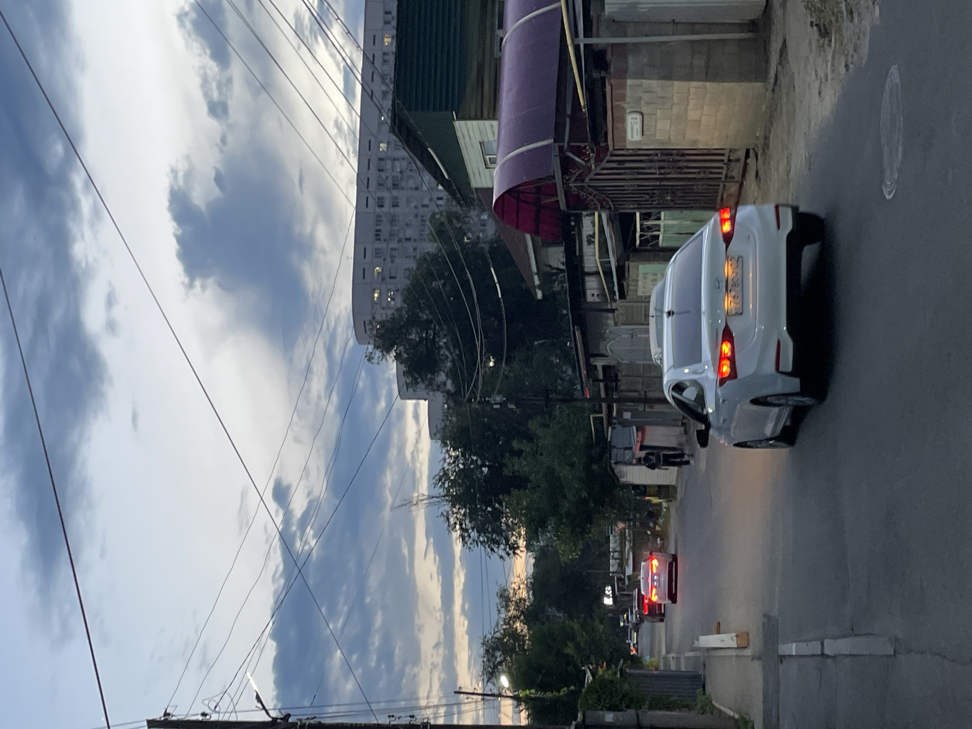 The image depicts a street scene in Almaty, Kazakhstan, at dusk. A white sedan is driving away from the camera, its brake lights illuminated. The car is on a paved road lined with low-rise buildings on the right and a sidewalk on the left. Overhead, power lines crisscross the sky. In the background, a large, multi-story apartment building is visible, its windows lit. The sky is filled with dramatic, cloudy formations, suggesting impending weather. A lone pedestrian is walking away from the camera on the sidewalk to the right. Other vehicles are visible further down the street, also with their lights on. The overall impression is one of a typical urban evening in Almaty.