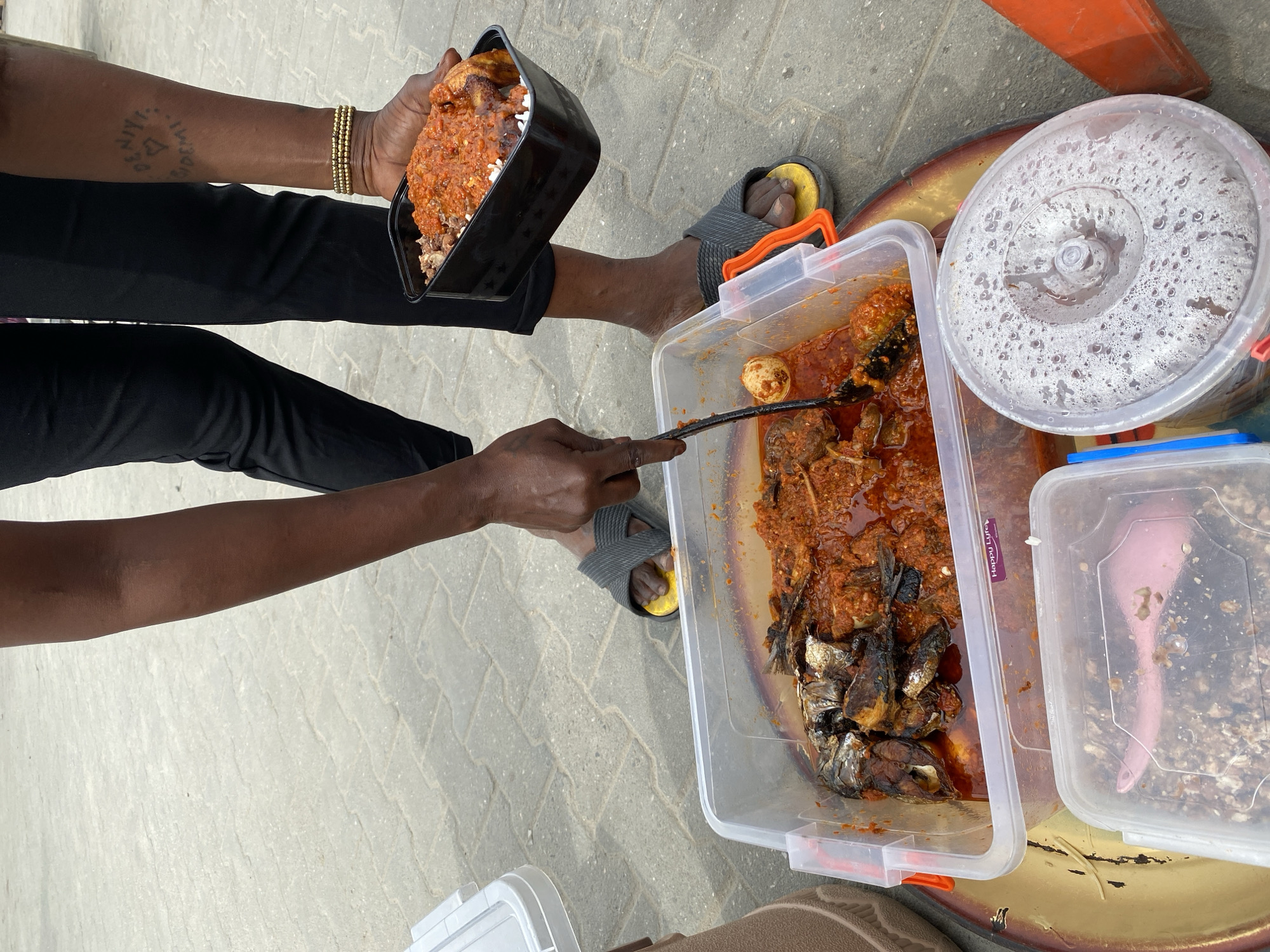 The image captures a street food scene, likely in Lagos, Nigeria, given the style of food preparation and environment. A person is serving food from a large, rectangular, transparent plastic container filled with a stew-like dish containing fish, eggs, and other ingredients. The stew is red in color, suggesting the use of tomatoes and spices. The person is using a long, dark-colored spoon to transfer the food into a black, rectangular food container being held by another person.
The person holding the container is wearing black pants and a gold bracelet on their wrist. Their arm has a tattoo. They are also wearing grey and yellow flip-flops.
Next to the large container of stew, there is a round plastic container with a lid covered in what appears to be condensation or water droplets. There is also a smaller, square plastic container with a lid to its right.
The scene is set outdoors on a paved surface made of grey paving stones. The overall impression is that of a bustling street food environment, with food being prepared and served directly to customers.