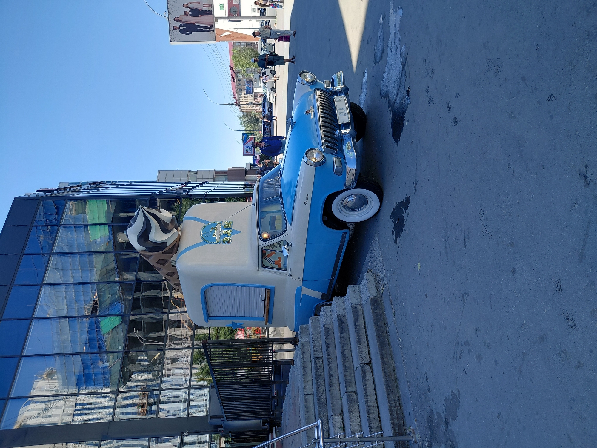 An antique blue and white ice cream truck is parked on a paved surface in front of a modern building with a glass facade. The truck has a large ice cream cone decoration on its roof. Several people are visible in the background, walking and standing near other vehicles on a street. A billboard with an advertisement featuring a family is also visible in the distance. The sky is clear and blue. The scene appears to be a street in a city.