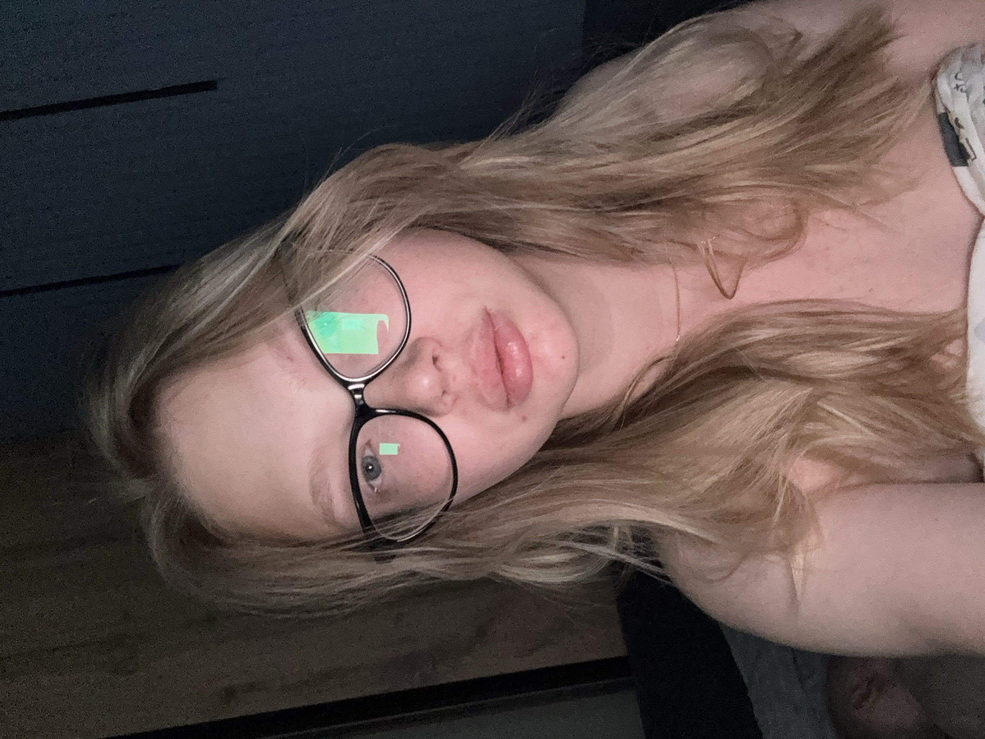 This is a medium shot selfie of a young woman with blonde hair and glasses. She is wearing a white garment and appears to be in a dimly lit room. The background is dark and out of focus, with some texture visible on the left side which could be a wooden surface, and a dark wall on the right. A faint reflection of a green rectangular object is visible on her glasses. The woman is looking directly at the camera. There is a blurry figure visible in the lower left corner of the image, possibly another person. The image does not provide specific details to determine its location in Chaikovsky, Russia.