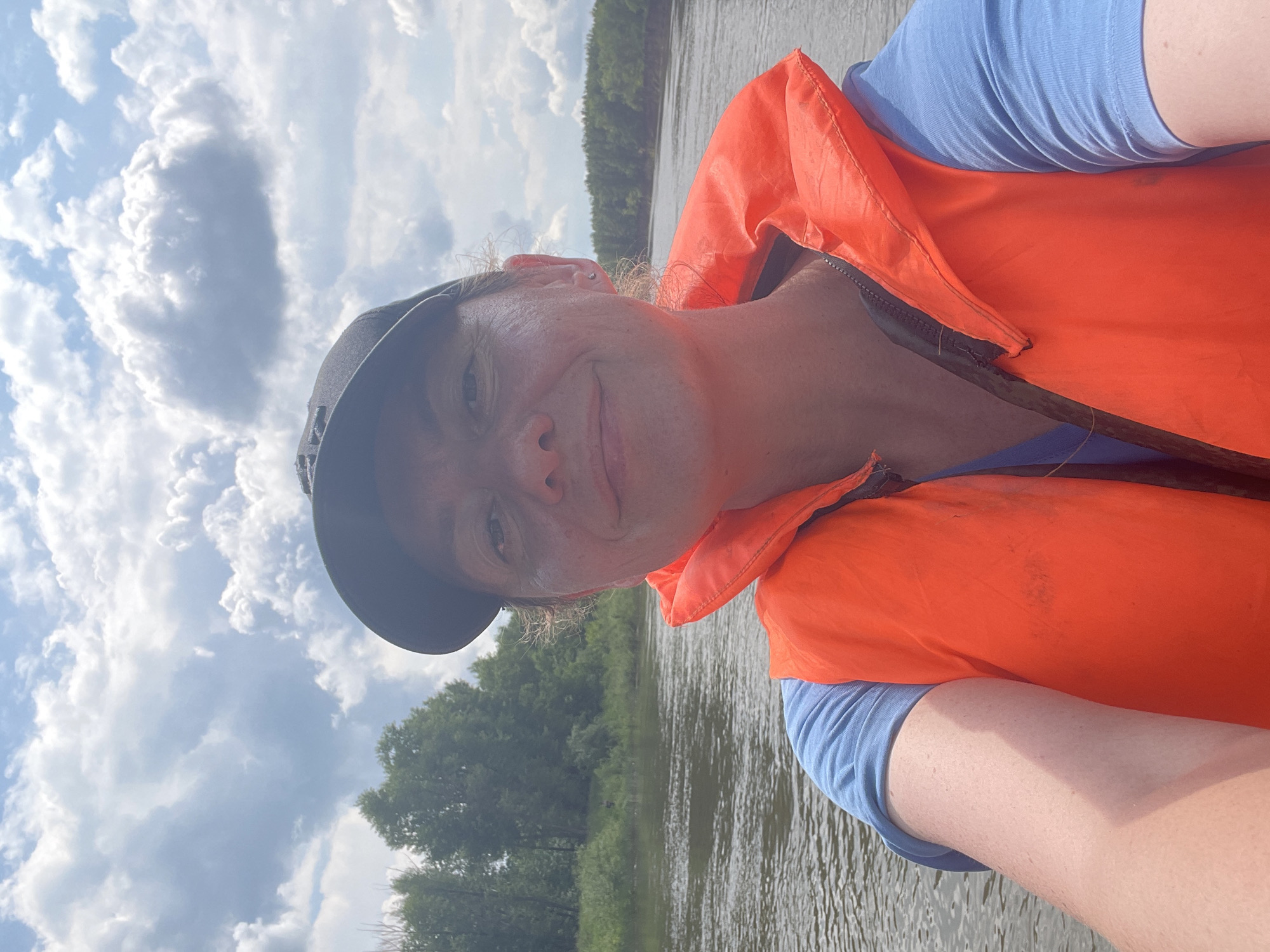 The image is a selfie taken by a woman wearing a blue t-shirt and an orange life vest, with a black baseball cap on her head. She is on a body of water, likely a river, with trees and vegetation lining the banks in the background. The sky is cloudy. The woman is smiling, and her face is partially visible. The scene appears to be outdoors, possibly in a natural or recreational area.