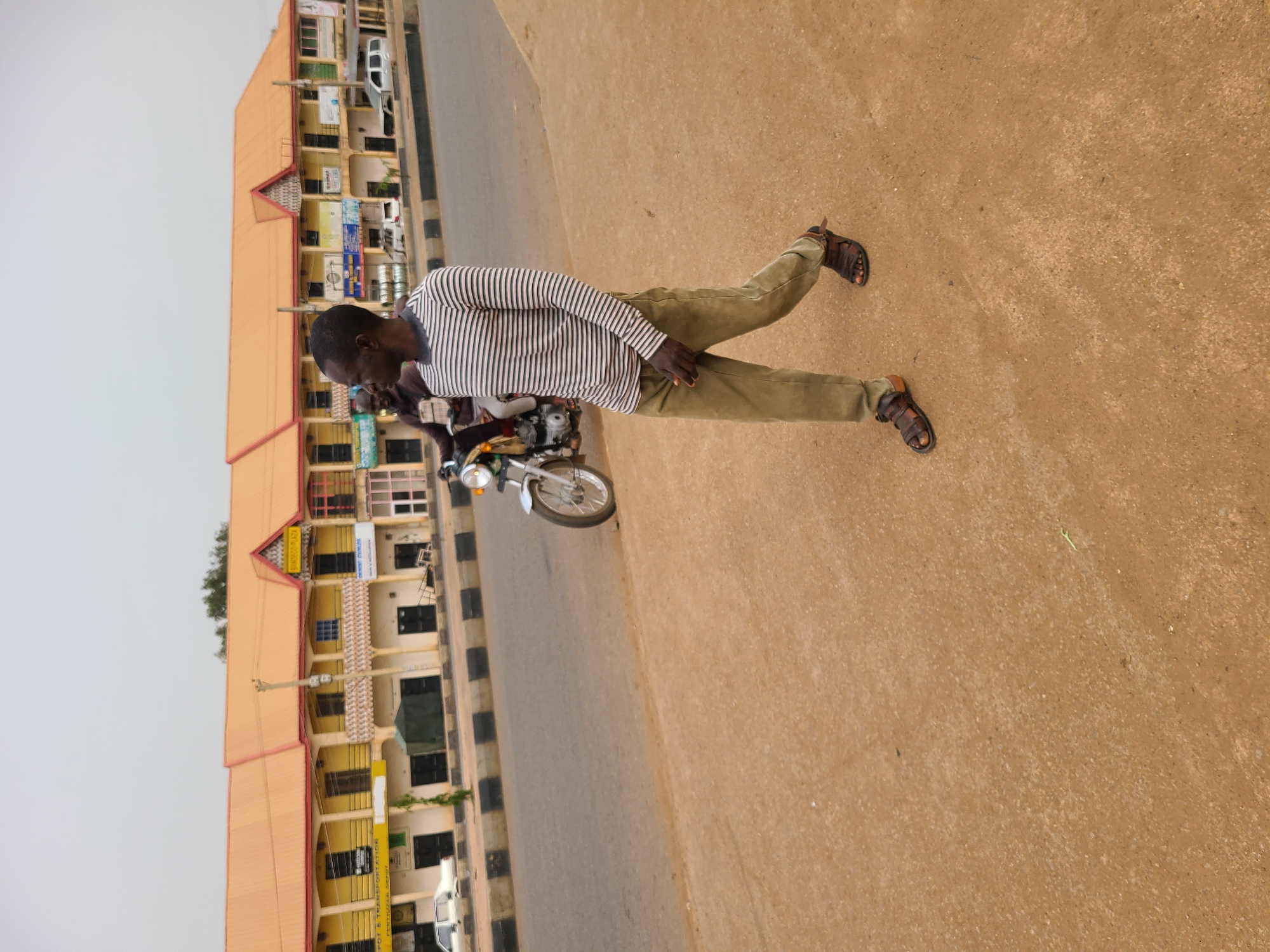 A man stands on a dirt shoulder next to a motorcycle parked on a road in Jos, Nigeria. The man is wearing a striped shirt, khaki pants, and sandals. The motorcycle is silver. A multi-story building stands on the other side of the road. The sky is overcast.