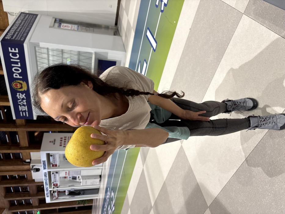 An indoor photograph captures a Caucasian female in mid-frame, bent slightly forward, holding a large, spherical yellow fruit to her nose as if smelling it. Her eyes are closed, and she has a slight smile. She wears a light-colored t-shirt, a light blue long-sleeved top tied around her waist, dark grey patterned leggings, and grey hiking-style boots with red laces. The floor consists of light and dark grey checkerboard tiles, traversed by a distinct green and blue striped lane with white markings. In the background, two white modular booths are visible. The booth on the left is labeled "INTELLIGENT SERVICE AREA" in English and Chinese. The booth on the right is a police station kiosk, featuring a prominent dark blue sign with "三亚公安" (Sanya Public Security) and "POLICE" in English, along with the Chinese police emblem. A smaller sign on the right side of the police kiosk mentions "Temporary Driving Permit for Foreigners". The scene is situated within a modern public facility, likely a transit or administrative building, in Tianya District, Sanya, China.