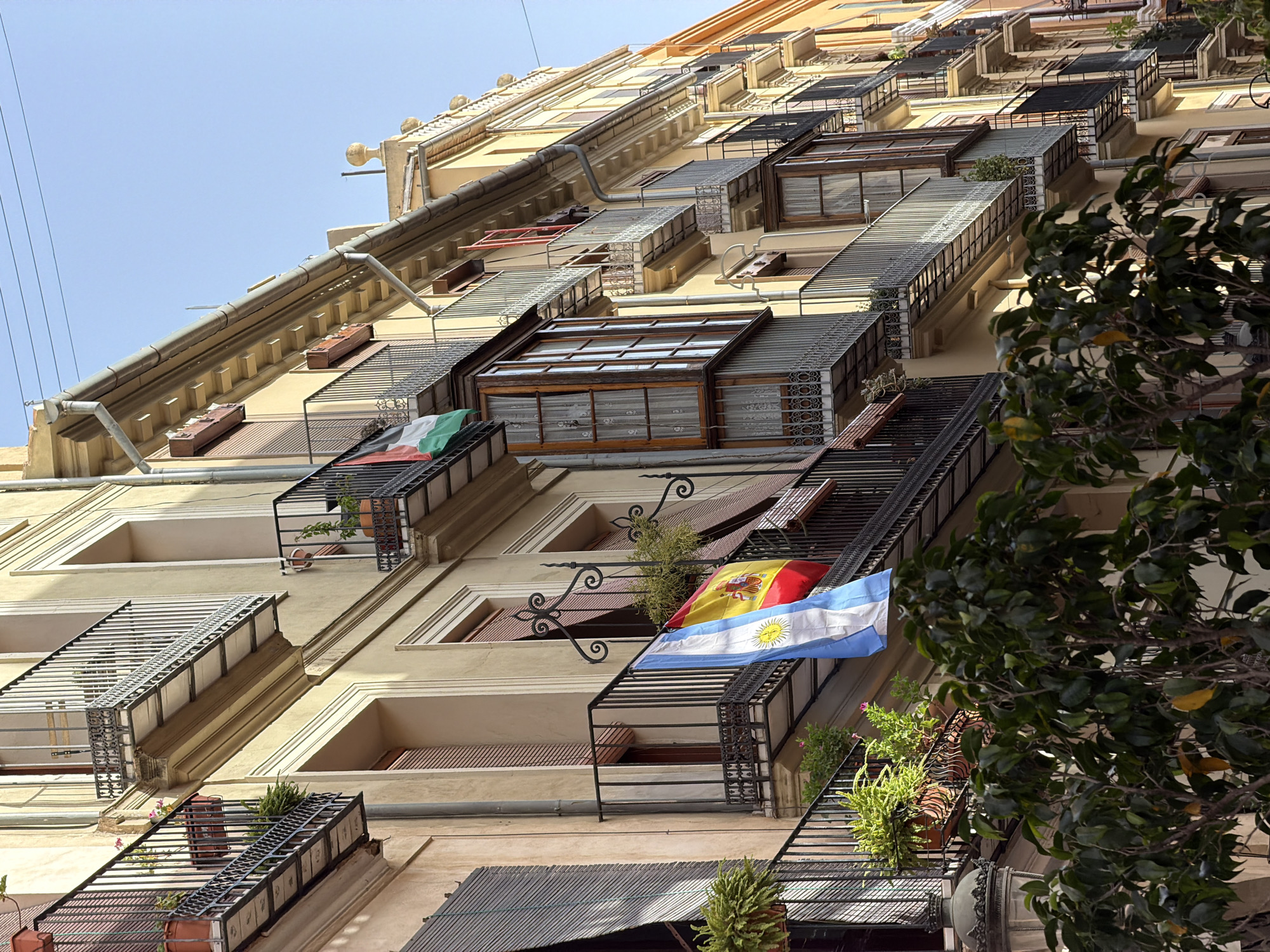 The image shows a low-angle, close-up view of a pale yellow building facade with multiple balconies. The building extends out of frame to the right and is in a state of shadow and light. There are several balconies with black metal railings. On the first visible balcony, from left to right, there is a large potted fern. On the next balcony, also from left to right, are plants in terra cotta pots and two flags hanging. The flag on the left is blue and white with a sun emblem in the center, which appears to be the flag of Argentina. To its right is a red and yellow flag with a crest, which appears to be the flag of Spain. Further to the right, there are more balconies with plants and awnings. The architecture suggests a Mediterranean or European urban setting. There are no people visible in the image.