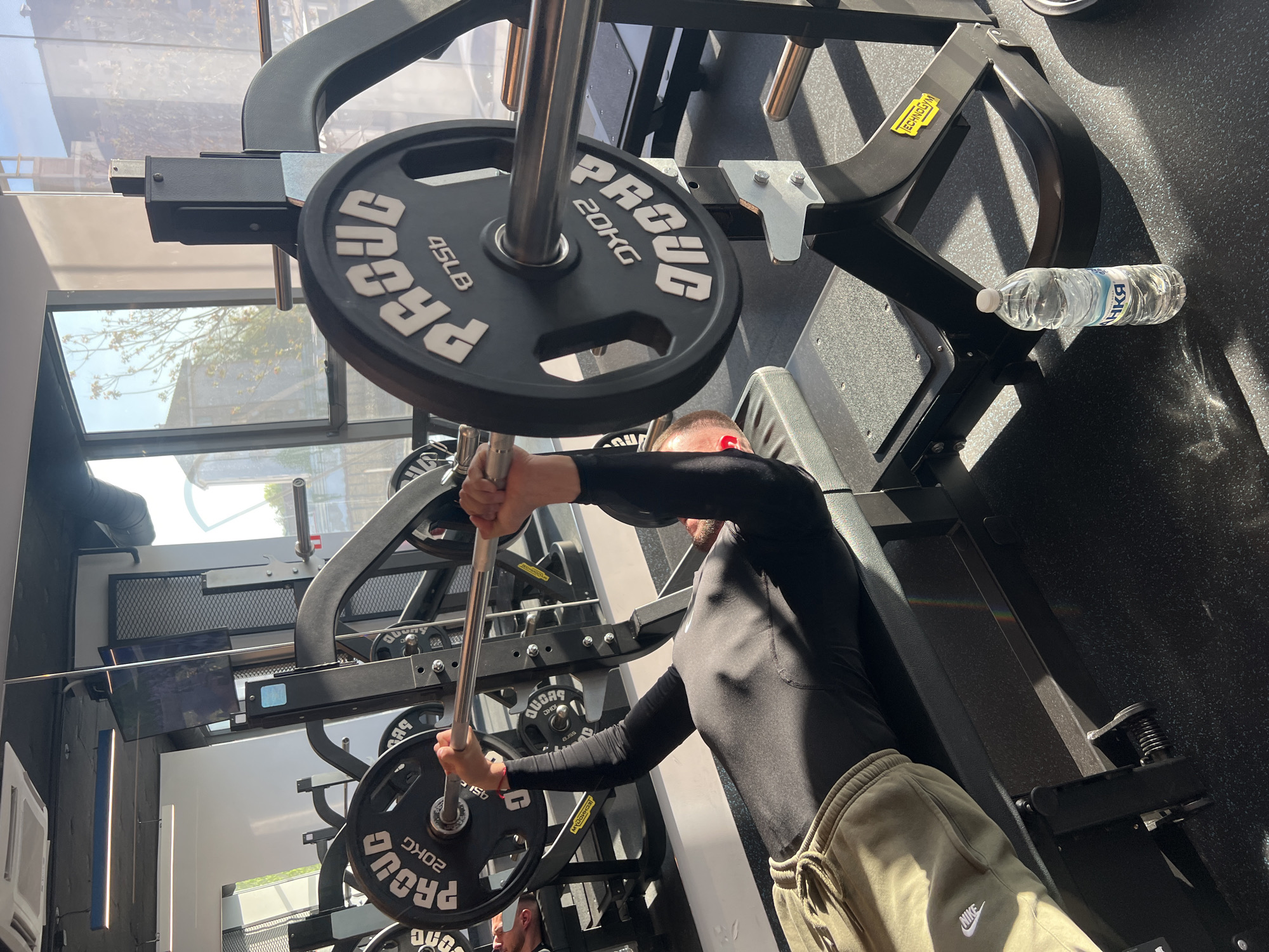 An indoor gym scene in Plovdiv, Bulgaria, captures a man engaged in weightlifting. The man, wearing a black long-sleeve top and olive green sweatpants with a visible "NIKE" logo, is standing at a large black gym machine, likely a leg press or hack squat, gripping a bar with both hands. He has short hair and appears to have a red earbud in his right ear.
The gym features modern equipment, including several black weight plates labeled "PROUD," with specifications "45LB" and "20KG" clearly visible. A yellow "TECHNOGYM" sticker is also noticeable on the machine frame. The floor is covered with black, textured gym flooring. Sunlight streams in through large windows, illuminating parts of the gym and creating distinct shadows, suggesting it is daytime. Outside the windows, trees and buildings are faintly visible. A plastic water bottle, with the text "АНКЯ" (ANKYA) on its label, rests on a part of the gym equipment. The overall setting is bright and spacious, indicative of a well-equipped fitness facility.