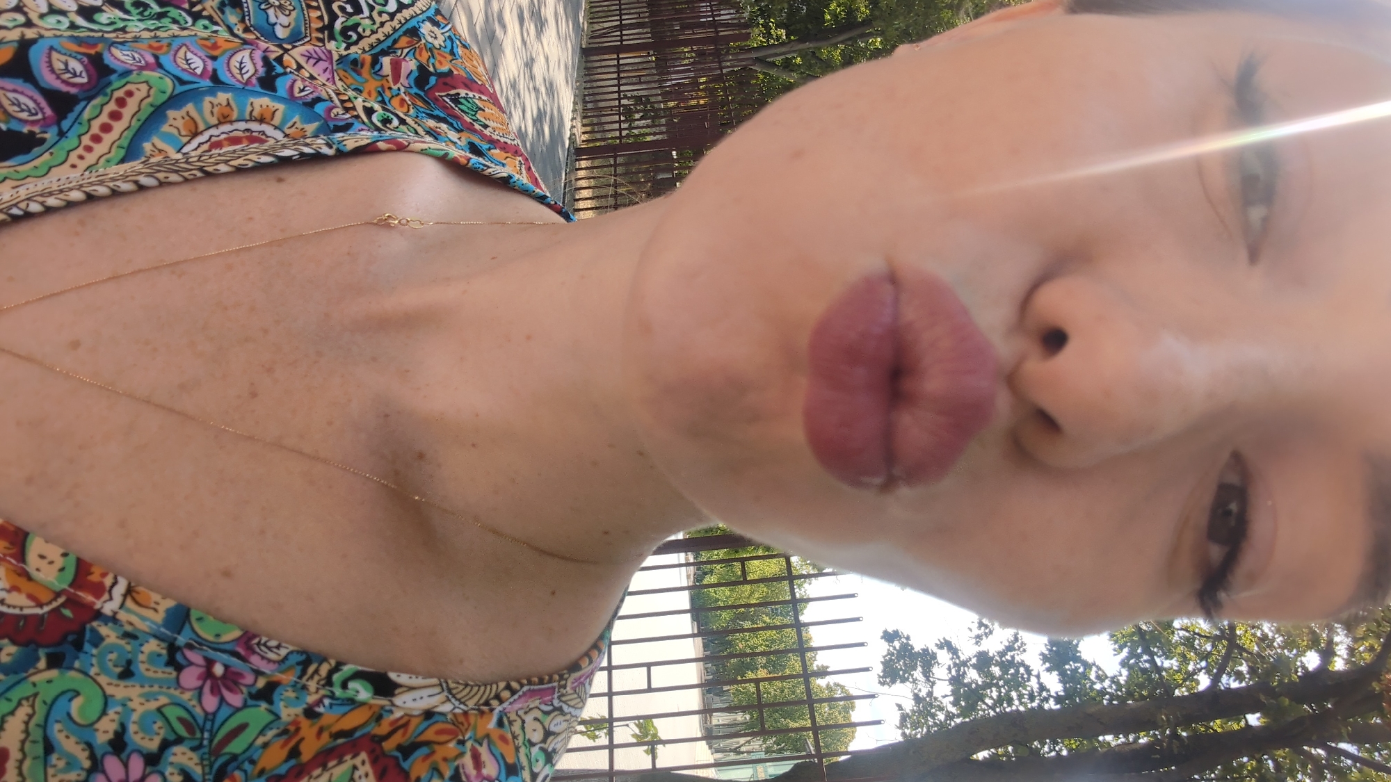 A close-up, waist-up, outdoor, selfie-style, full-color photograph features a woman posing with puckered lips. She has fair skin with freckles, dark eyebrows, and is wearing dark eyeliner. The woman's hair is short and brown, framing her face. Her lips are painted with a matte, pink lipstick. She is wearing a vibrant, patterned top with floral and paisley designs in shades of blue, green, yellow, pink, and red. A delicate gold necklace is visible around her neck. The background consists of a blurred outdoor environment with a metal fence, trees, and what appears to be a sidewalk or paved area. The lighting suggests it is daytime, with a strong light source causing a lens flare on the left side of the image. There are no other individuals or distinct objects in the immediate vicinity of the subject. The location is not identifiable as Chișinău, Moldova.