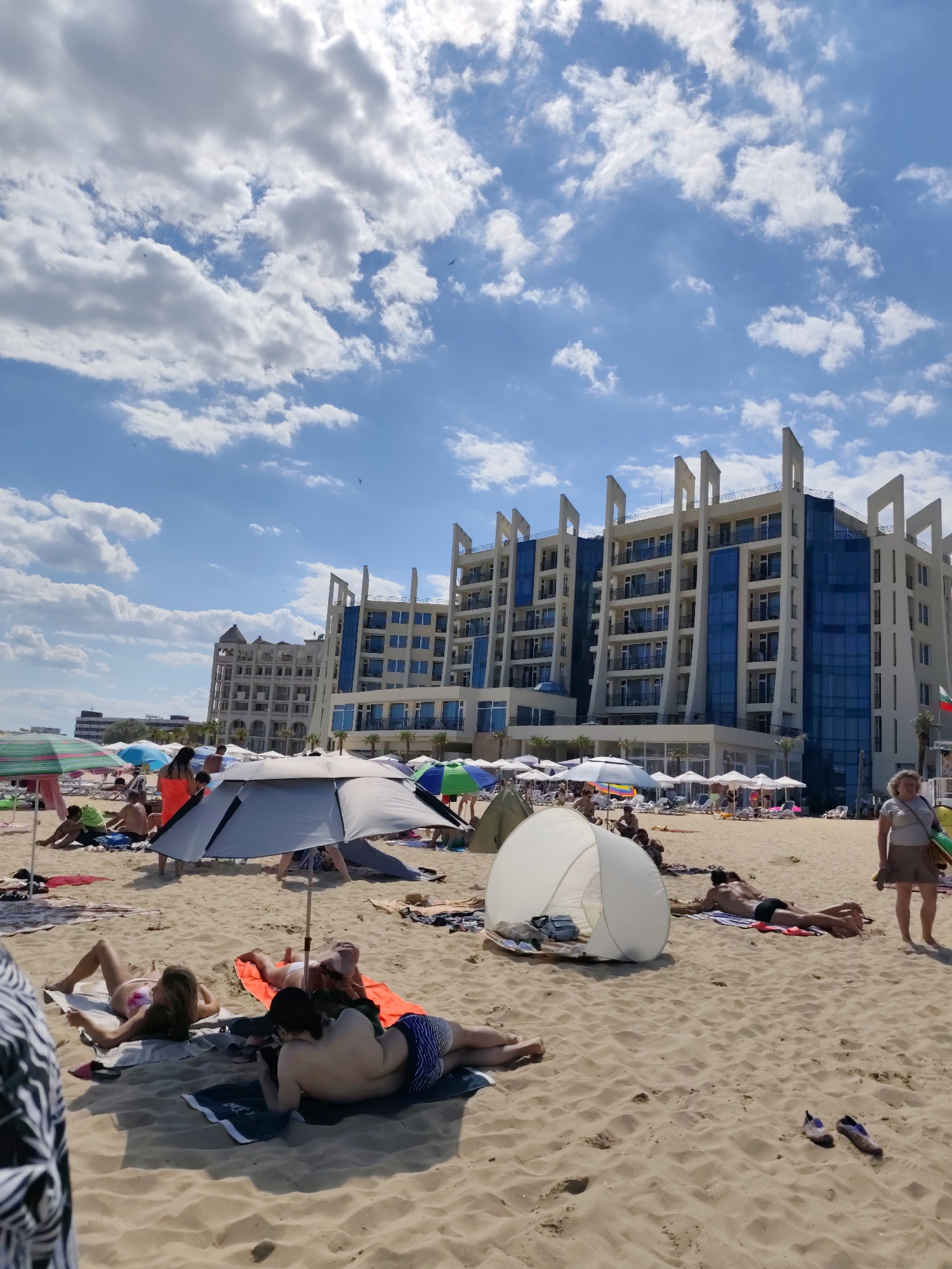 This image depicts a crowded beach scene in Nesebar, Bulgaria, with a large, modern hotel in the background. The beach is covered in sand, with numerous beach umbrellas and tents providing shade for sunbathers. People are scattered across the beach, many lying on towels or beach chairs, some reading or using their phones. The sea is not visible in this frame, but the presence of a beach strongly implies its proximity. The sky is blue with scattered white clouds. The hotel itself is a multi-story building with a contemporary architectural design, featuring glass balconies and white facade elements. In the foreground, several individuals are sunbathing on towels. The overall impression is a busy summer day at a popular seaside resort.