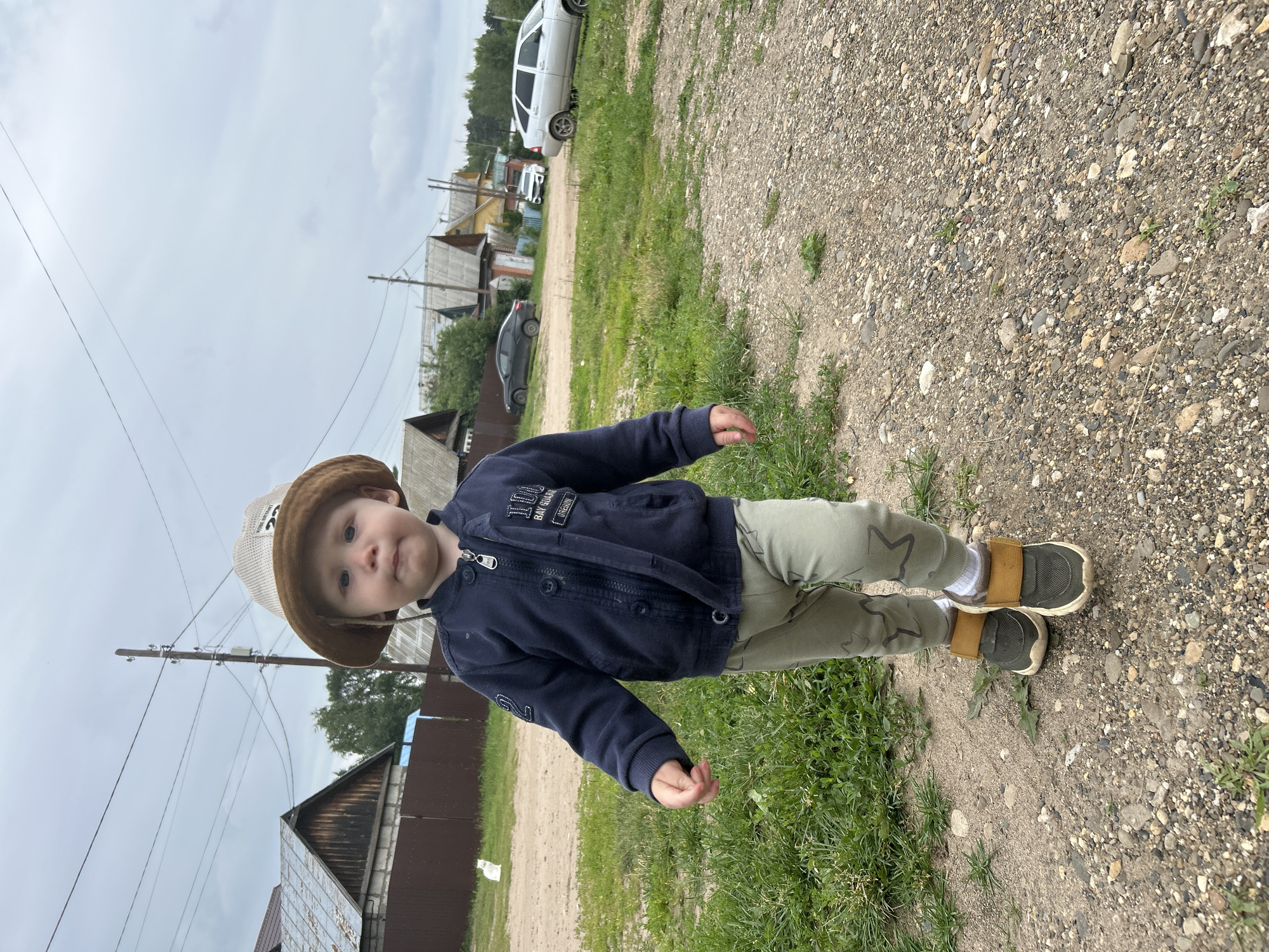 A young child, likely a toddler, is standing on a gravel path outdoors. The child is wearing a brown bucket hat, a dark blue zip-up jacket with some patches and text, light green patterned pants, white socks, and dark shoes with tan accents. The child is looking towards the camera with a neutral expression.
The setting appears to be a rural or suburban area with houses visible in the background, some with wooden siding. There are also power lines and a utility pole. A white car and a dark car are parked in the distance, along with other buildings and trees. The ground is covered in gravel and patches of green grass. The sky is overcast and appears to be a daytime scene.
The text on the child's jacket includes "100," "BAY GUARD," and "OCEAN." There is also a patch with a stylized "2". The hat has some text, but it is not clearly legible.
The image is oriented vertically, with the child in the foreground and the background receding. The overall impression is of a casual outdoor moment.