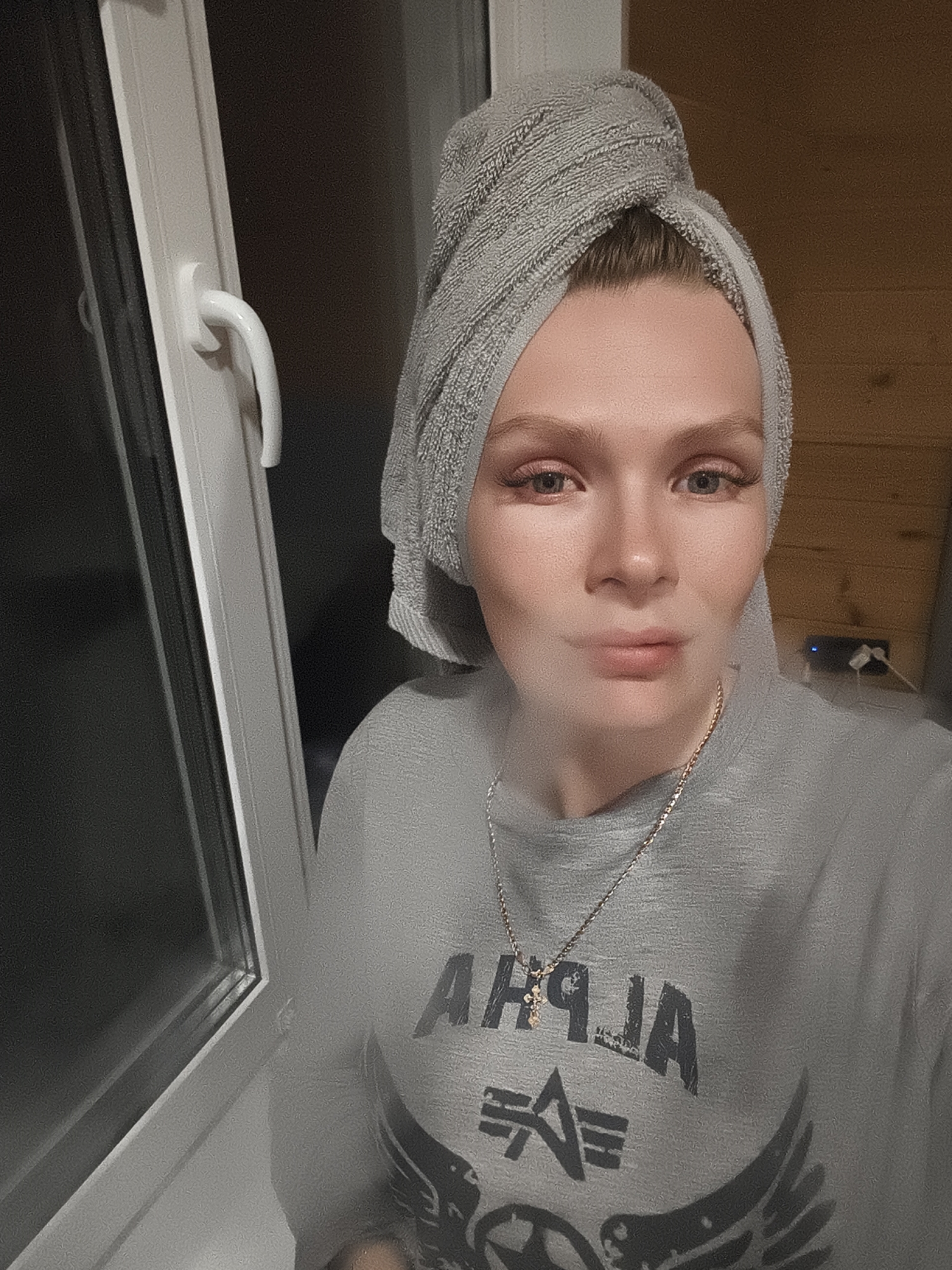 A selfie of a fair-skinned woman with blue eyes and a gray towel wrapped around her head. She is wearing a gray T-shirt with the word "ALPHA" in black letters. She is also wearing a gold necklace with a cross pendant. She appears to be smoking or exhaling vapor.
She is standing next to a white window frame.
There is a wooden wall visible in the background with a power outlet and charging cables.
The location is Русский Вожой, Russia.